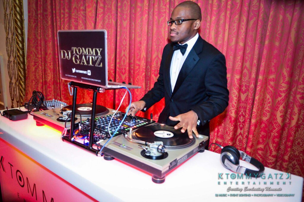 A DJ in a tuxedo operates turntables behind a DJ booth with "Tommy Gatz Entertainment" branding on the front.
