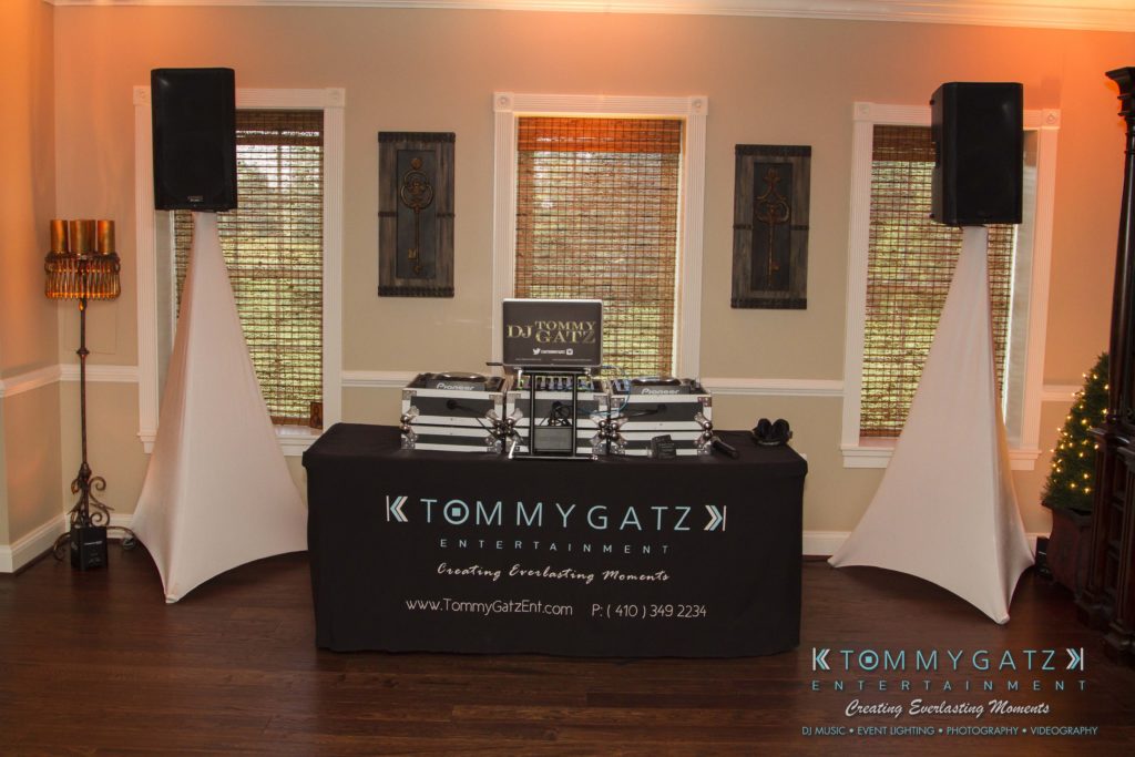 02-1024x683 - TOMMY GATZ ENTERTAINMENT DJ booth setup with speakers, lighting, and promotional signage from Tommy Gatz Entertainment in a room with wooden floors and blinds on windows.
