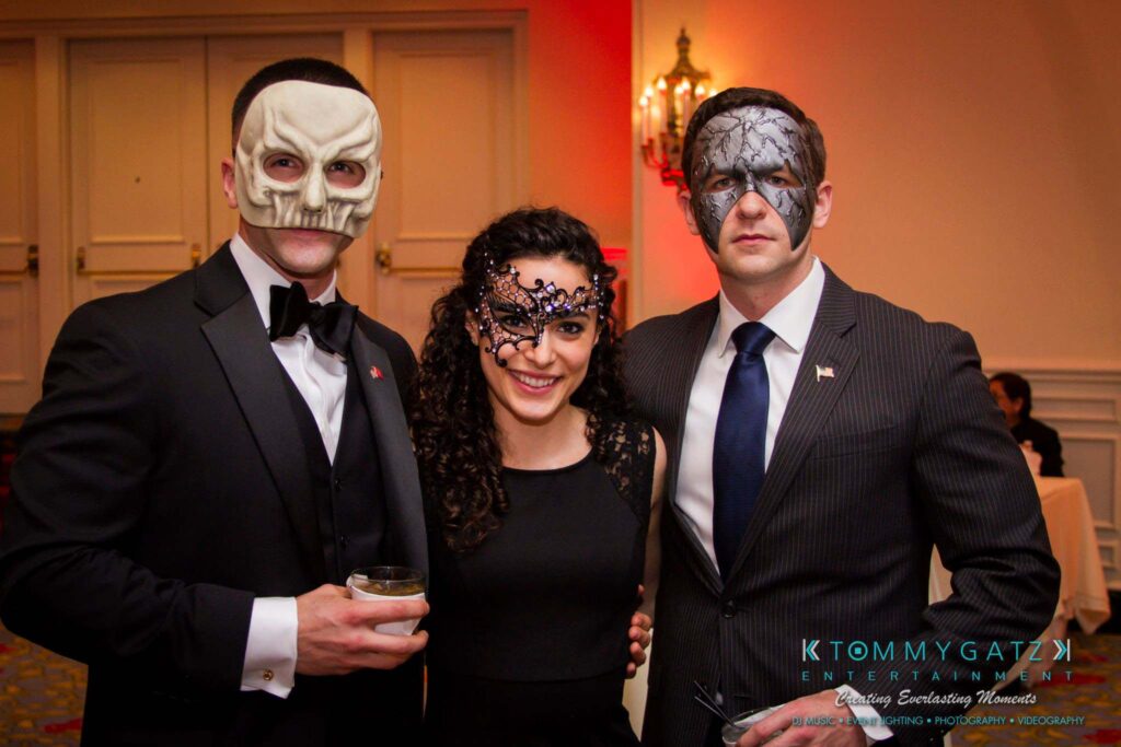 Three people pose for a photo wearing formal attire and decorative masks at a masquerade-themed event.