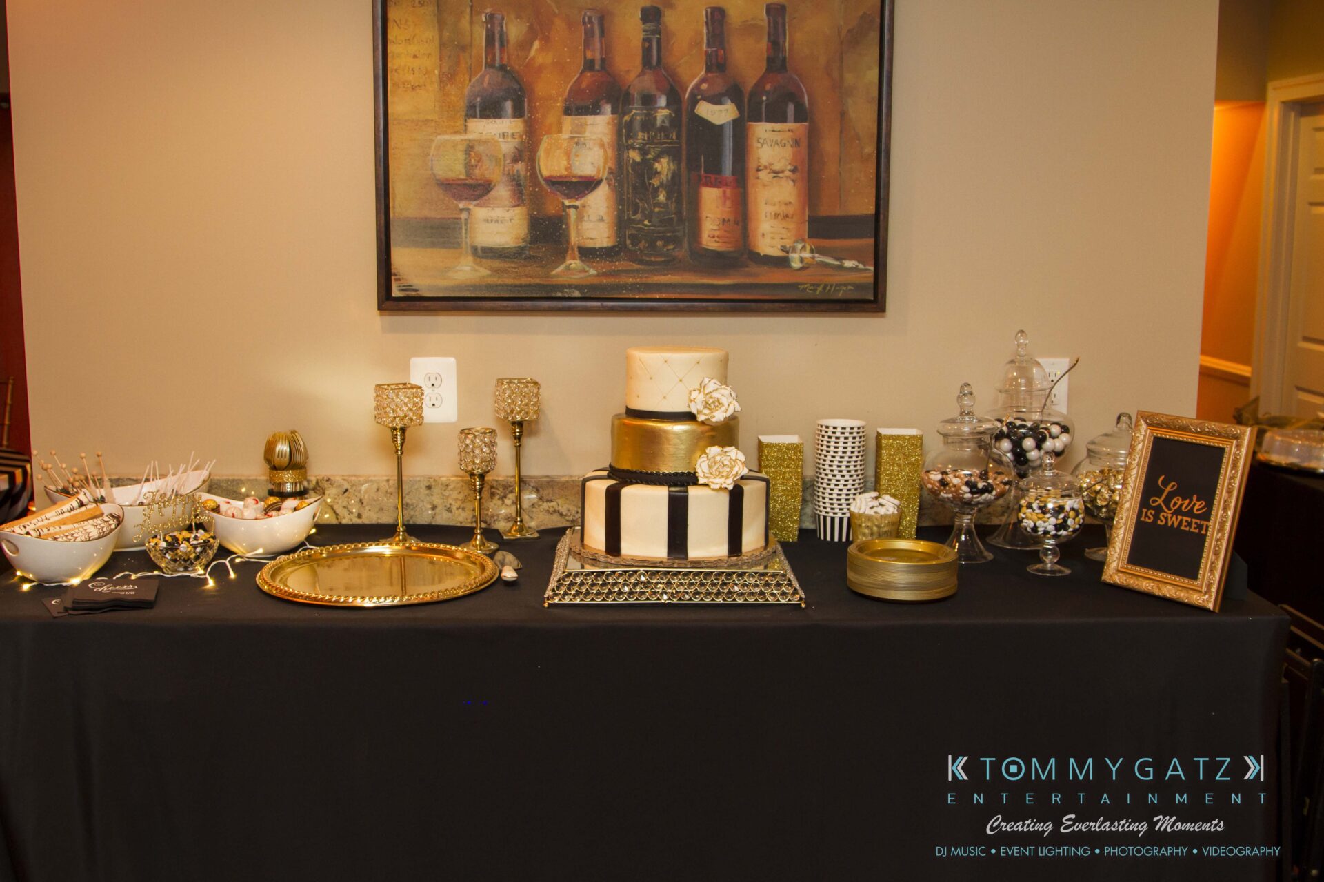 03 - TOMMY GATZ ENTERTAINMENT A dessert table featuring a tiered cake, assorted candies in jars, a gold tray, and a framed "Love is Sweet" sign, set against a backdrop of a wine-themed painting.