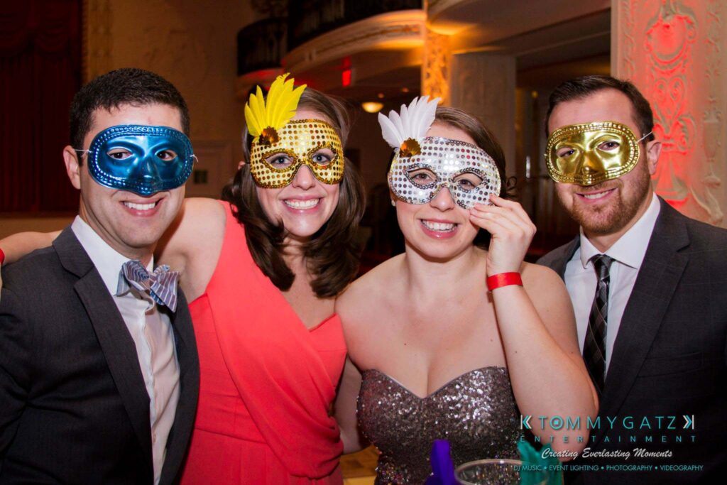 Four people wearing colorful masquerade masks and formal attire pose together in a festive indoor setting.