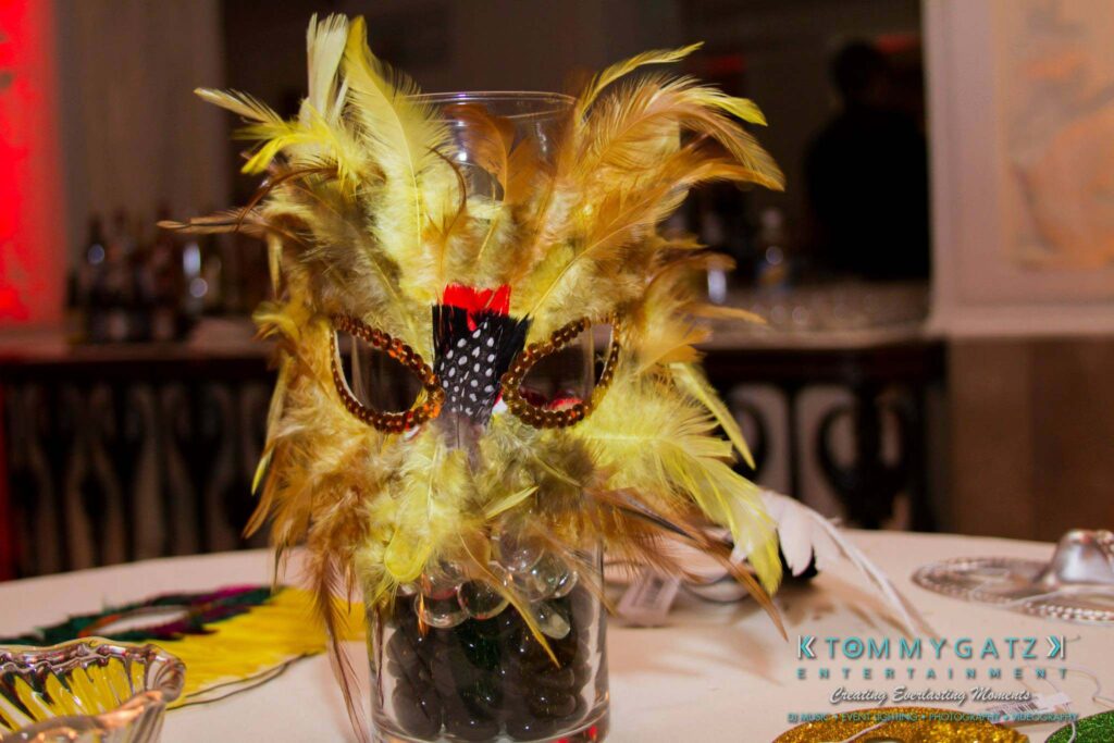 Yellow feather masquerade mask on a glass vase centerpiece with a logo in the background.
