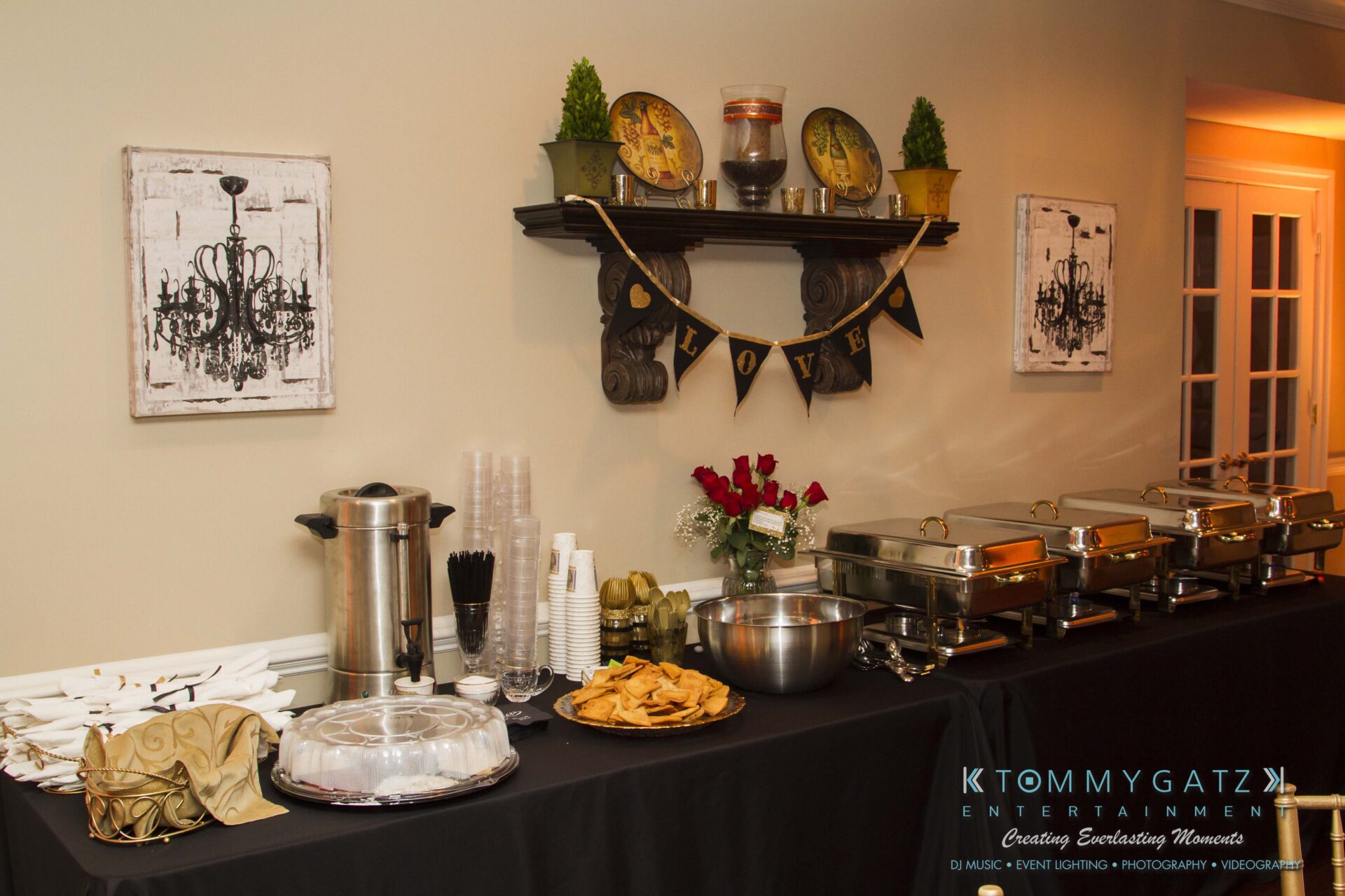05 - TOMMY GATZ ENTERTAINMENT Buffet table with covered dishes, chafing dishes, plates, cups, and a coffee urn. Decor includes wall clocks, a "LOVE" banner, and flowers.