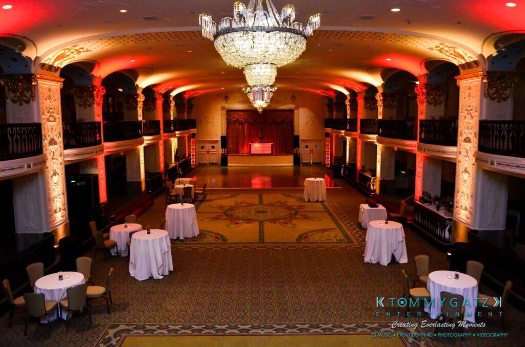 Elegant ballroom with ornate chandeliers and uplighting, featuring round tables with white tablecloths arranged around a central dance floor.