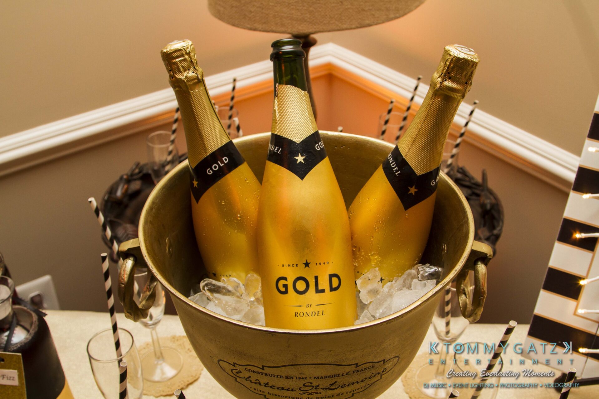 06 1 - TOMMY GATZ ENTERTAINMENT Three bottles of gold-colored sparkling wine in an ice-filled bucket on a table surrounded by black and white straws.