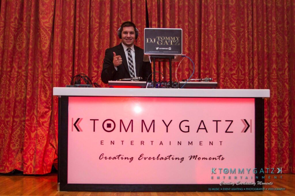 A person in a suit stands behind a DJ booth with the label "Tommy Gatz Entertainment" and gives a thumbs-up.