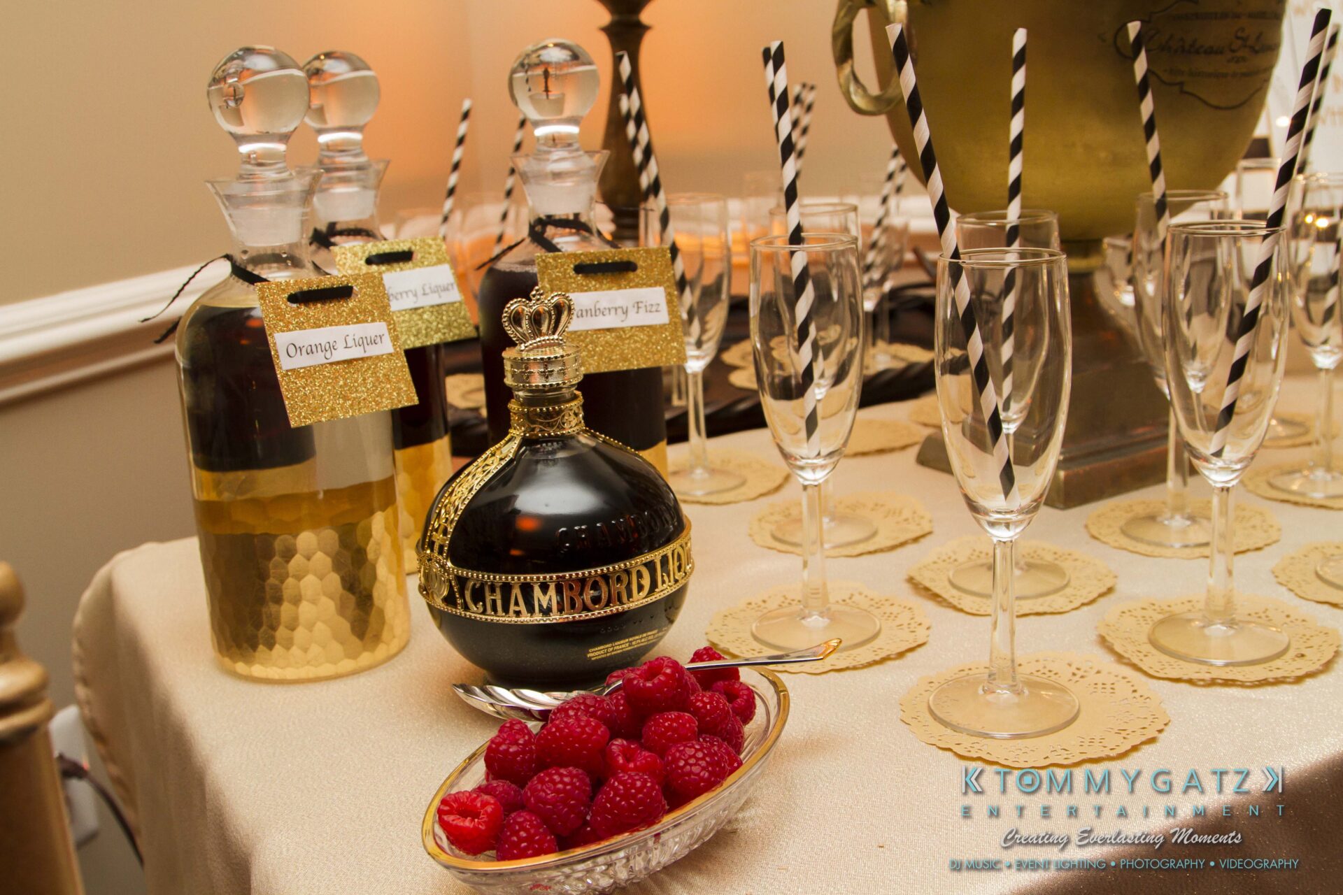 07 1 - TOMMY GATZ ENTERTAINMENT Table with Chambord bottle, two liqueurs, champagne flutes, striped straws, and a plate of raspberries.