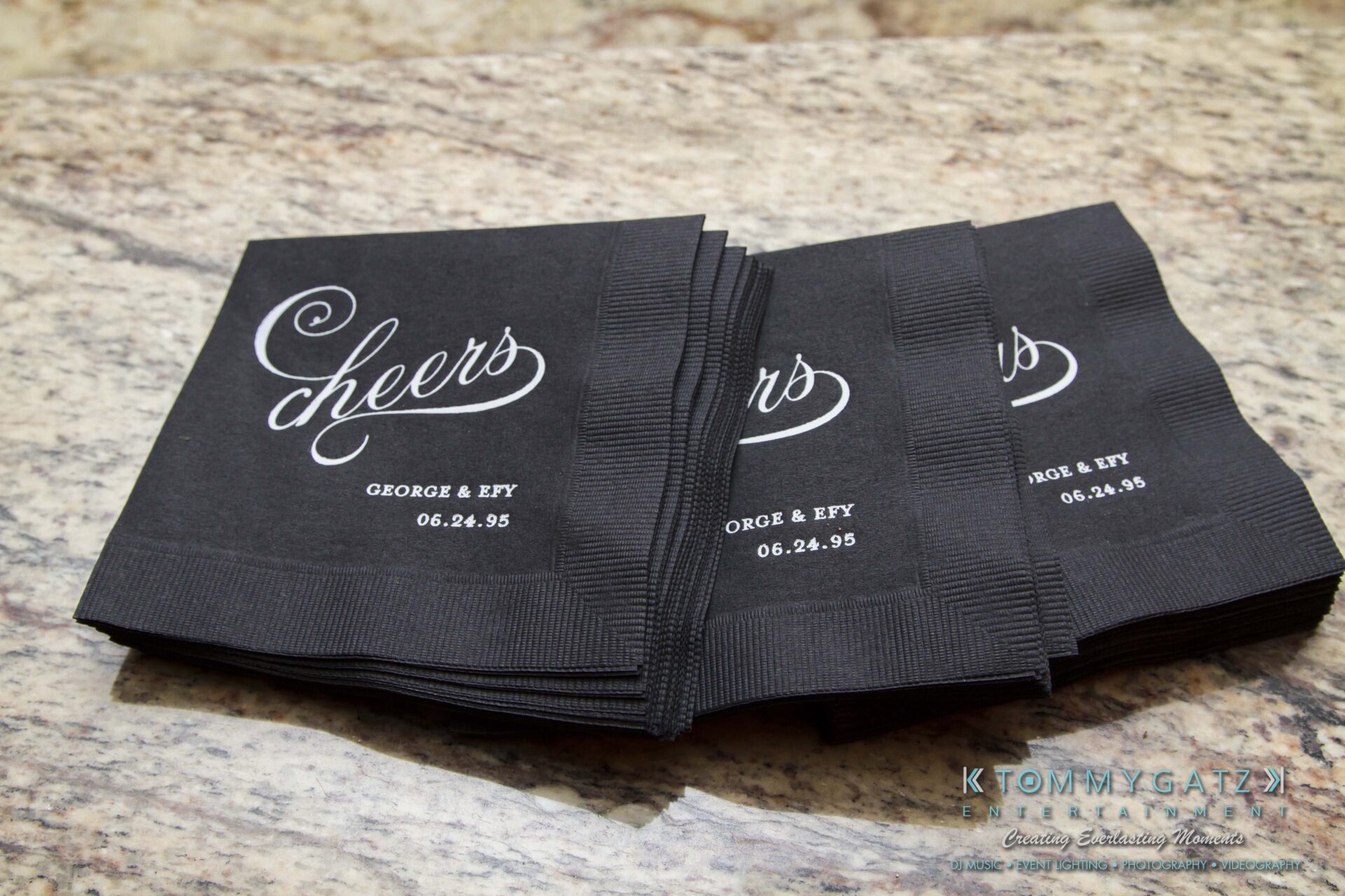 08 1 - TOMMY GATZ ENTERTAINMENT Stack of black napkins with "cheers" and "George & Efy 06.24.95" printed on them, placed on a stone surface.
