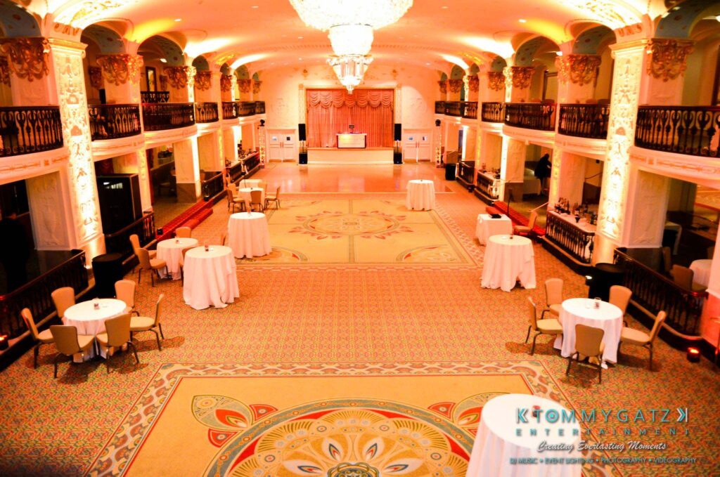 A spacious ballroom with ornate decor features round tables and chairs, balcony railings, and a chandelier. A DJ booth is set up at the far end.