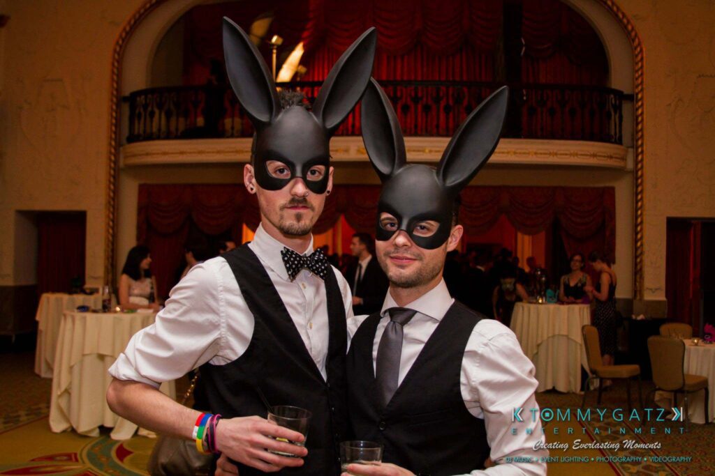 Two individuals wearing rabbit ear masks and formal attire pose indoors at an event. Tables with tablecloths and people are visible in the background.