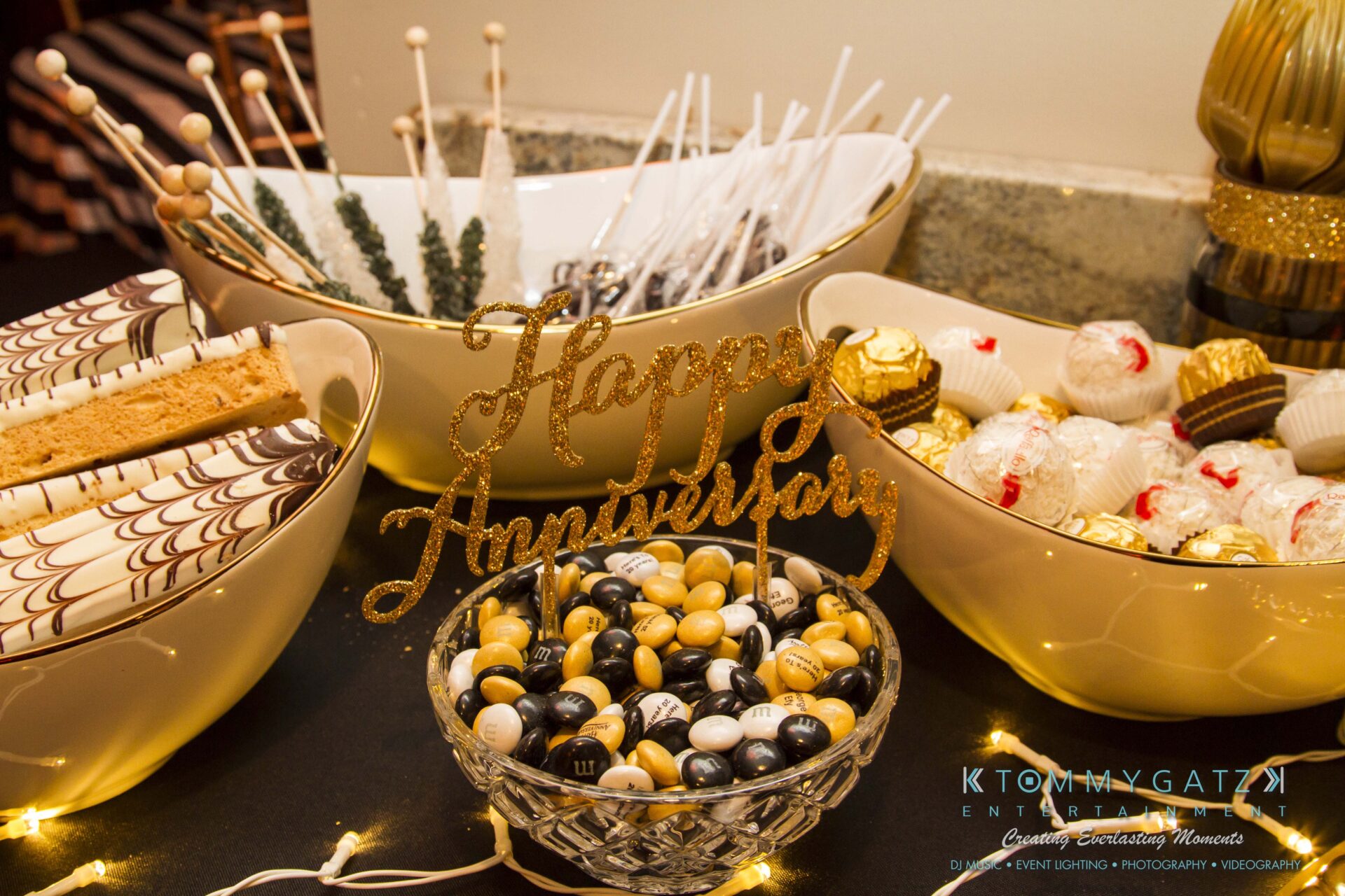 10 1 - TOMMY GATZ ENTERTAINMENT Dessert table with various sweets, including cake pops, chocolate truffles, cookies, and a bowl of black, white, and gold candies with a "Happy Anniversary" sign.