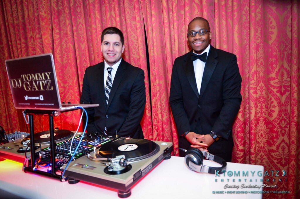 Two DJs in formal attire stand behind a DJ setup with turntables and a laptop, against a red curtain backdrop.