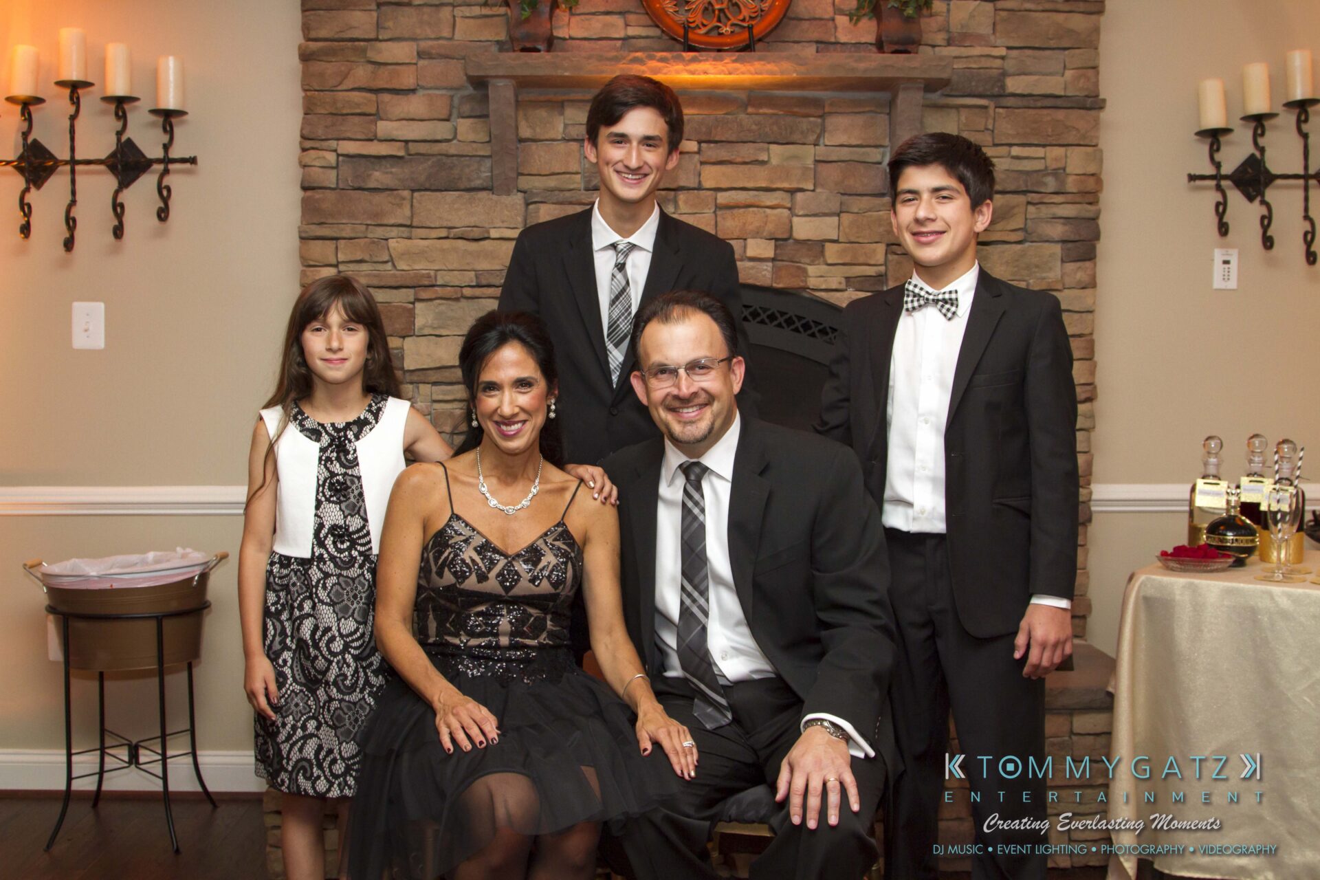 17 - TOMMY GATZ ENTERTAINMENT A family group photo with three boys, a girl, and a woman and man dressed formally, posing together in front of a stone fireplace.