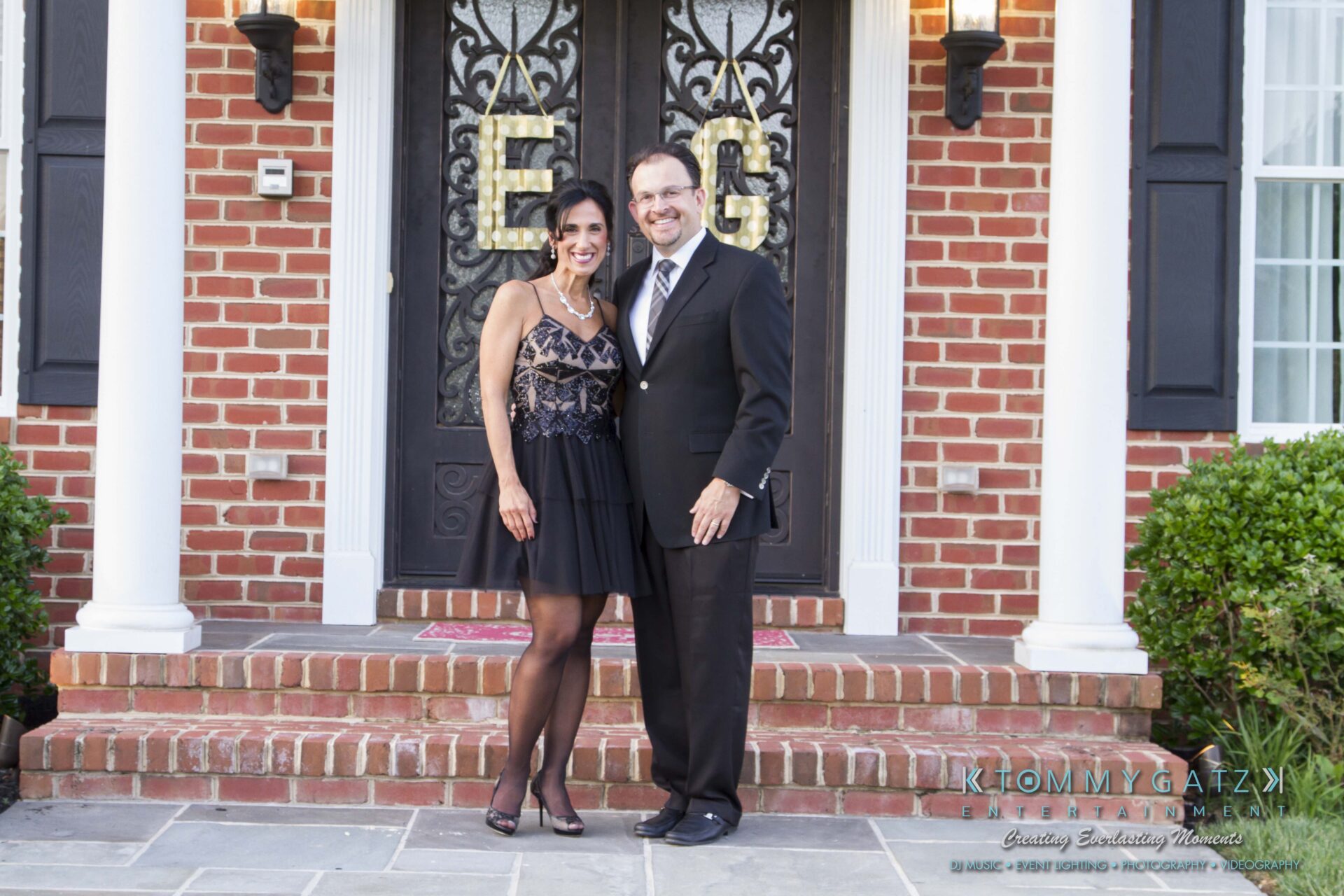 24 - TOMMY GATZ ENTERTAINMENT A couple in formal attire stands smiling in front of a red brick house with a black door.