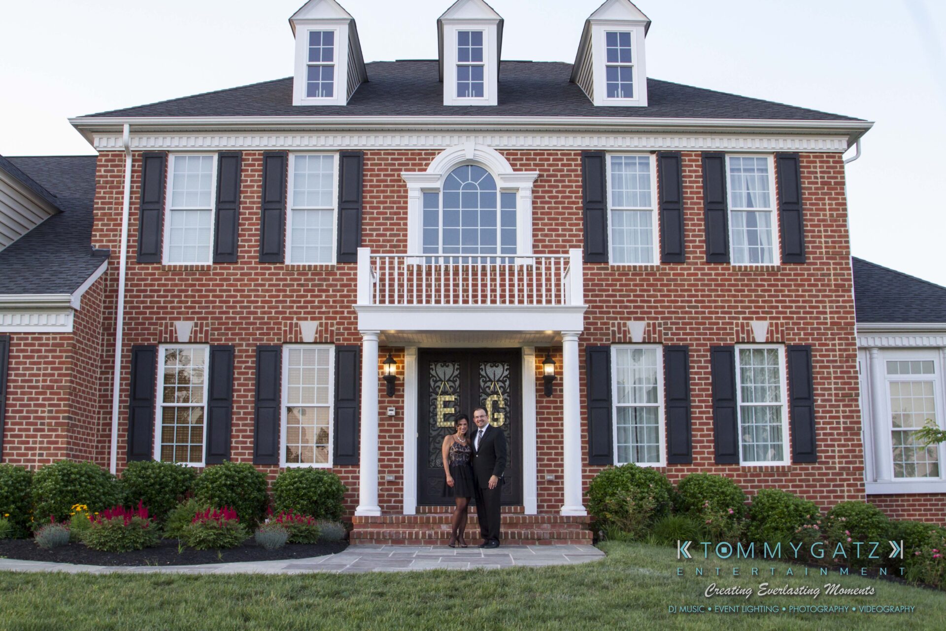 25 - TOMMY GATZ ENTERTAINMENT A man and woman stand in front of a large brick house with white trim and a manicured lawn.