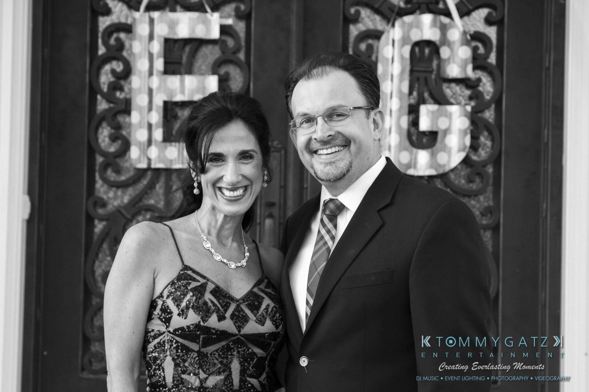 27 - TOMMY GATZ ENTERTAINMENT A man and a woman smiling in front of a decorative door with the letters E and G. The man is wearing a suit and tie, and the woman is in a patterned dress.