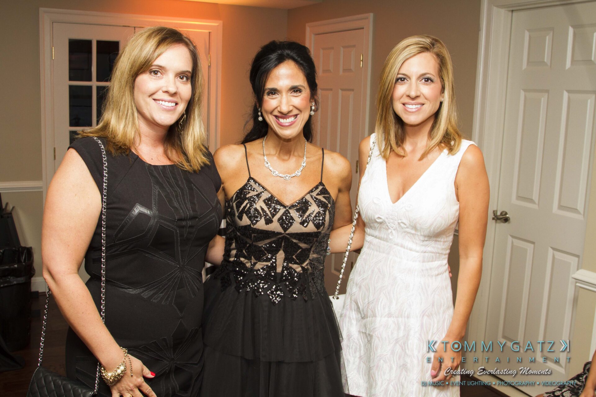 29 - TOMMY GATZ ENTERTAINMENT Three women in elegant dresses pose together at an indoor event with light-colored walls and closed doors in the background.