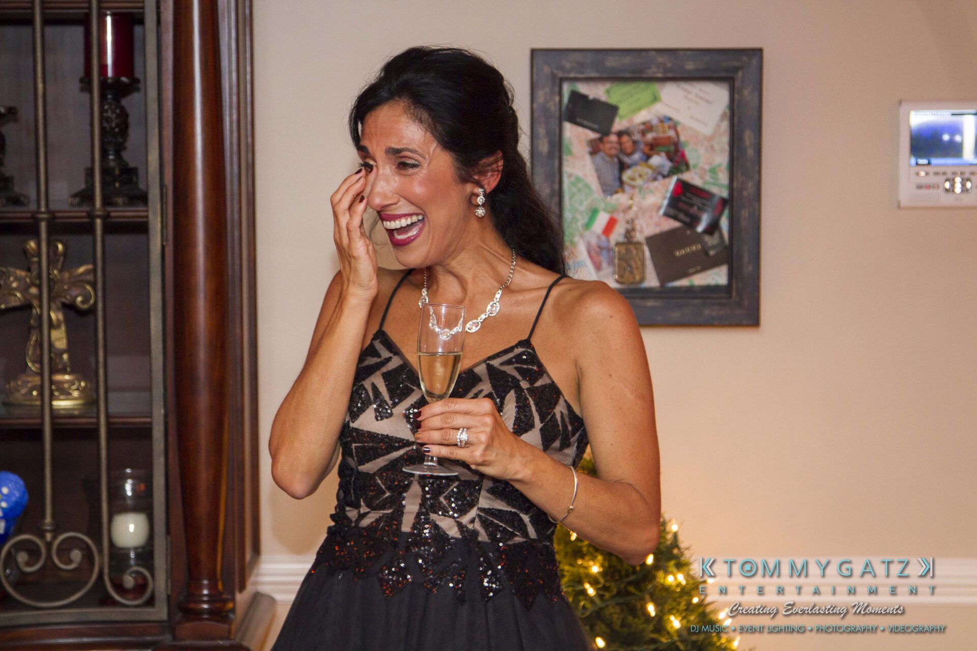 35 - TOMMY GATZ ENTERTAINMENT A woman in a black dress holds a champagne glass and wipes her eye, smiling in front of a shelf and framed photos.