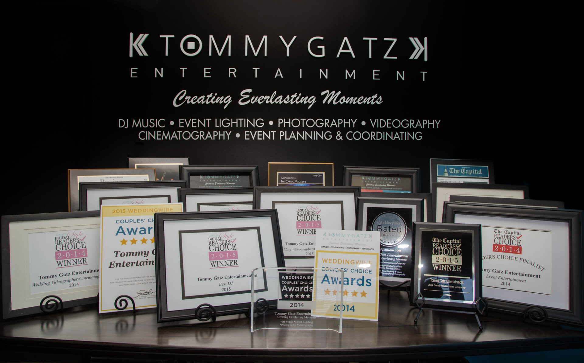 53 - TOMMY GATZ ENTERTAINMENT A collection of framed awards and certificates displayed on a table, with the words "Tommy Gatz Entertainment" on the wall behind them.
