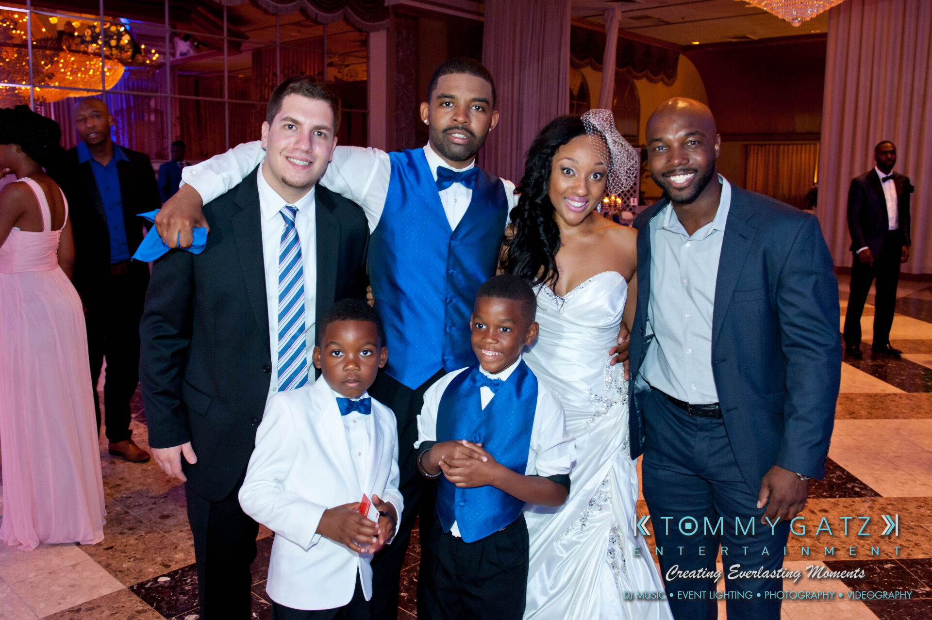 57 - TOMMY GATZ ENTERTAINMENT A group of five adults and two children, dressed in formal attire, pose together at a wedding reception.