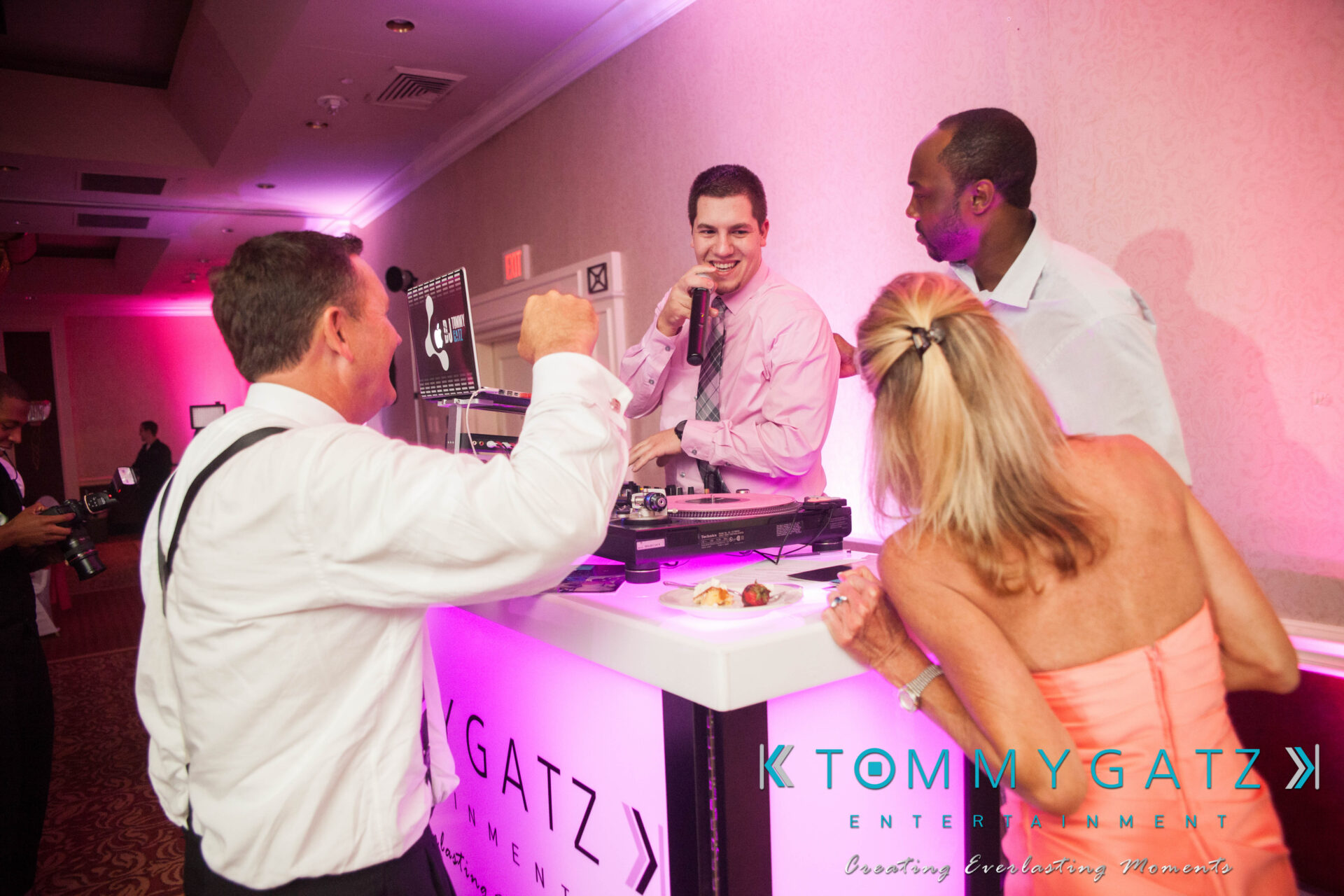 59 - TOMMY GATZ ENTERTAINMENT People enjoying an event with a DJ playing music at a booth, two men talking nearby, and a woman with a plate of food. Pink lighting adds ambiance to the room.
