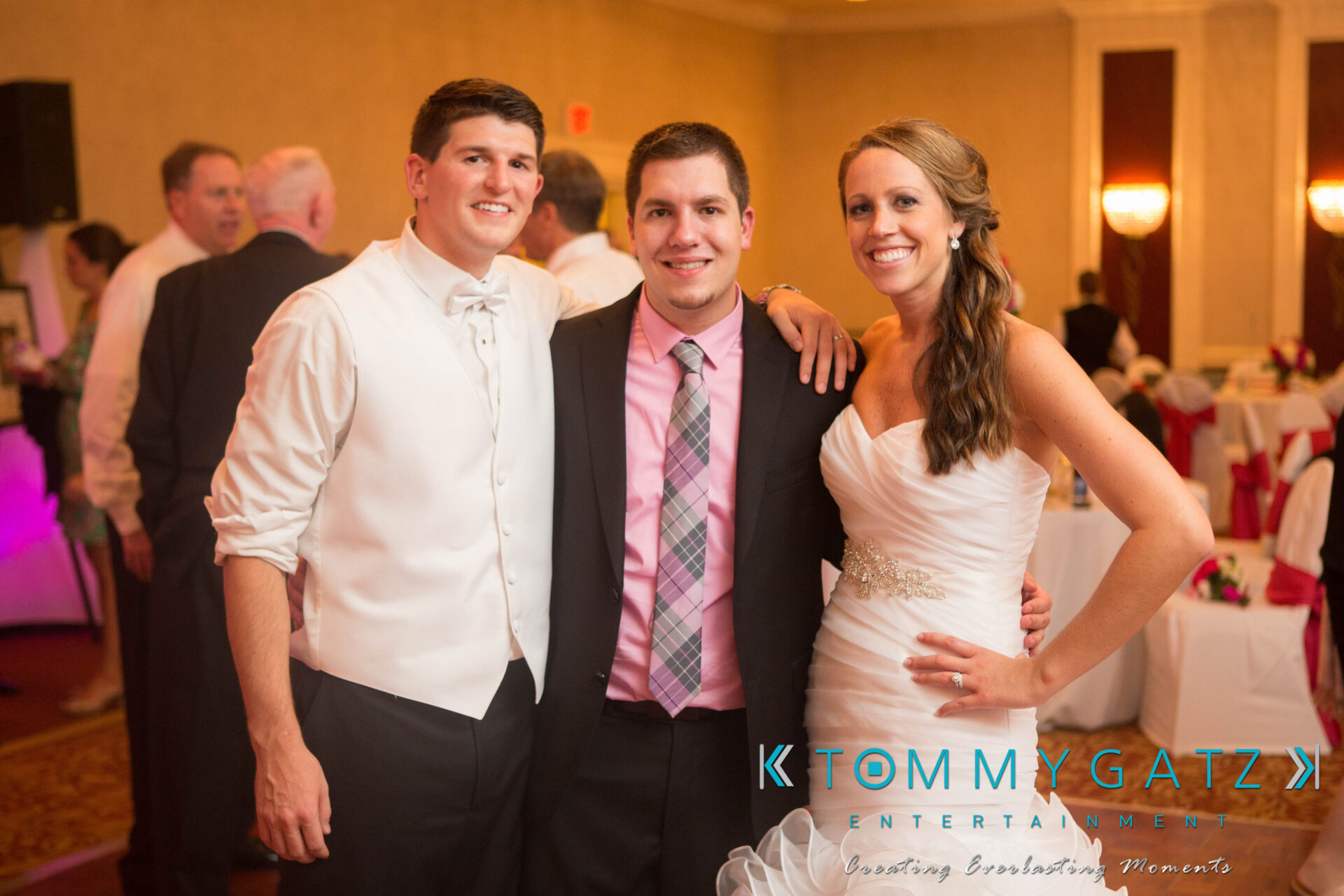 60-1 - TOMMY GATZ ENTERTAINMENT Three people pose together indoors at an event, with one wearing a wedding dress and the others dressed in formal attire.