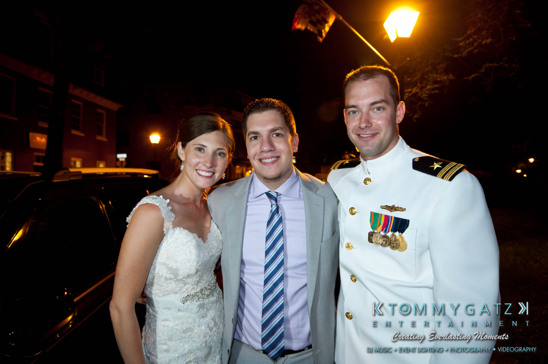 61 - TOMMY GATZ ENTERTAINMENT Three people standing together at night; a woman in a white dress, a man in a suit, and another man in a military uniform. They are smiling near a warmly lit street lamp.