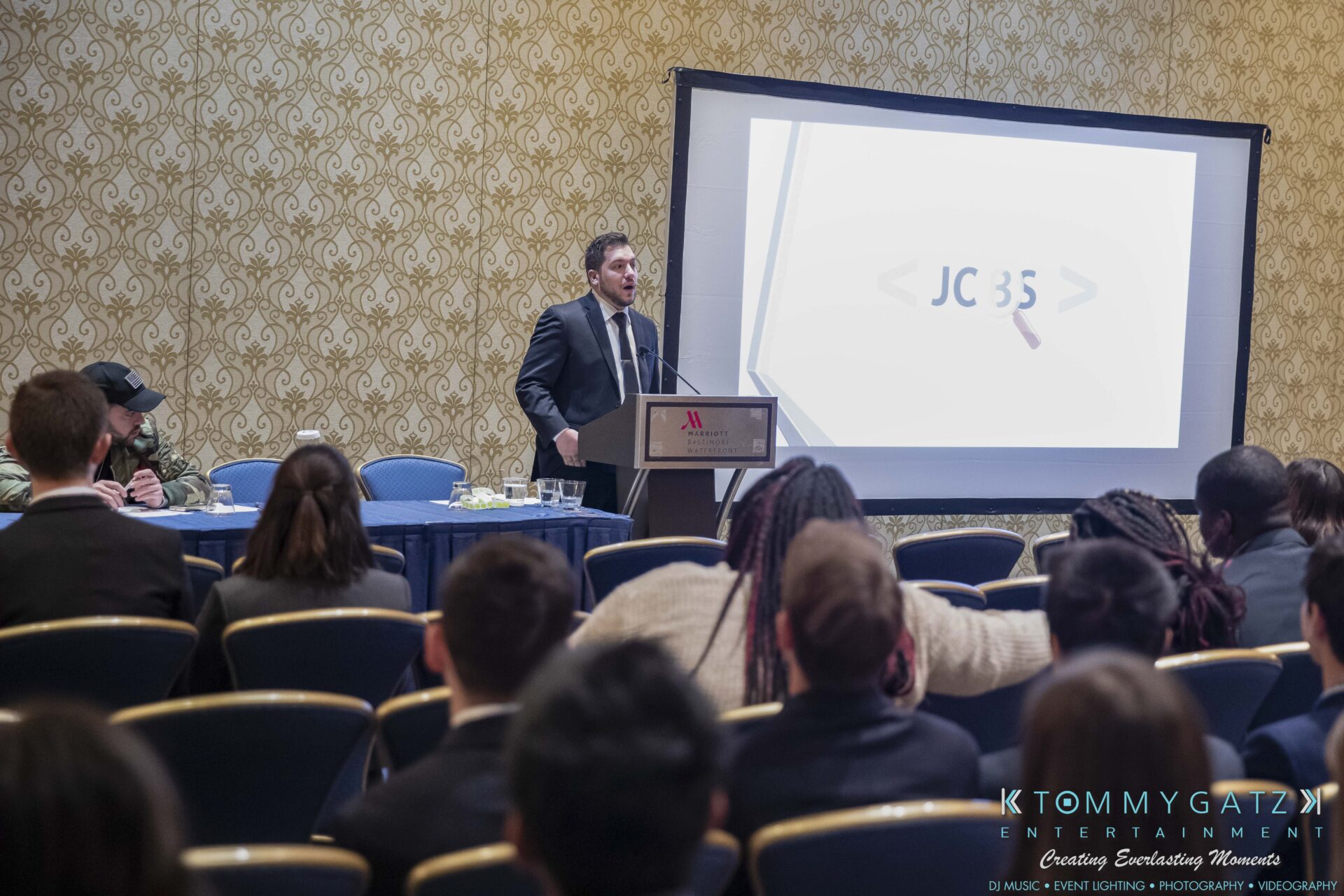 DECA-2018-Low-Res-WM-10 - TOMMY GATZ ENTERTAINMENT Person speaking at a podium in front of an audience, with a presentation slide displaying "JC35" on a screen in a conference room.