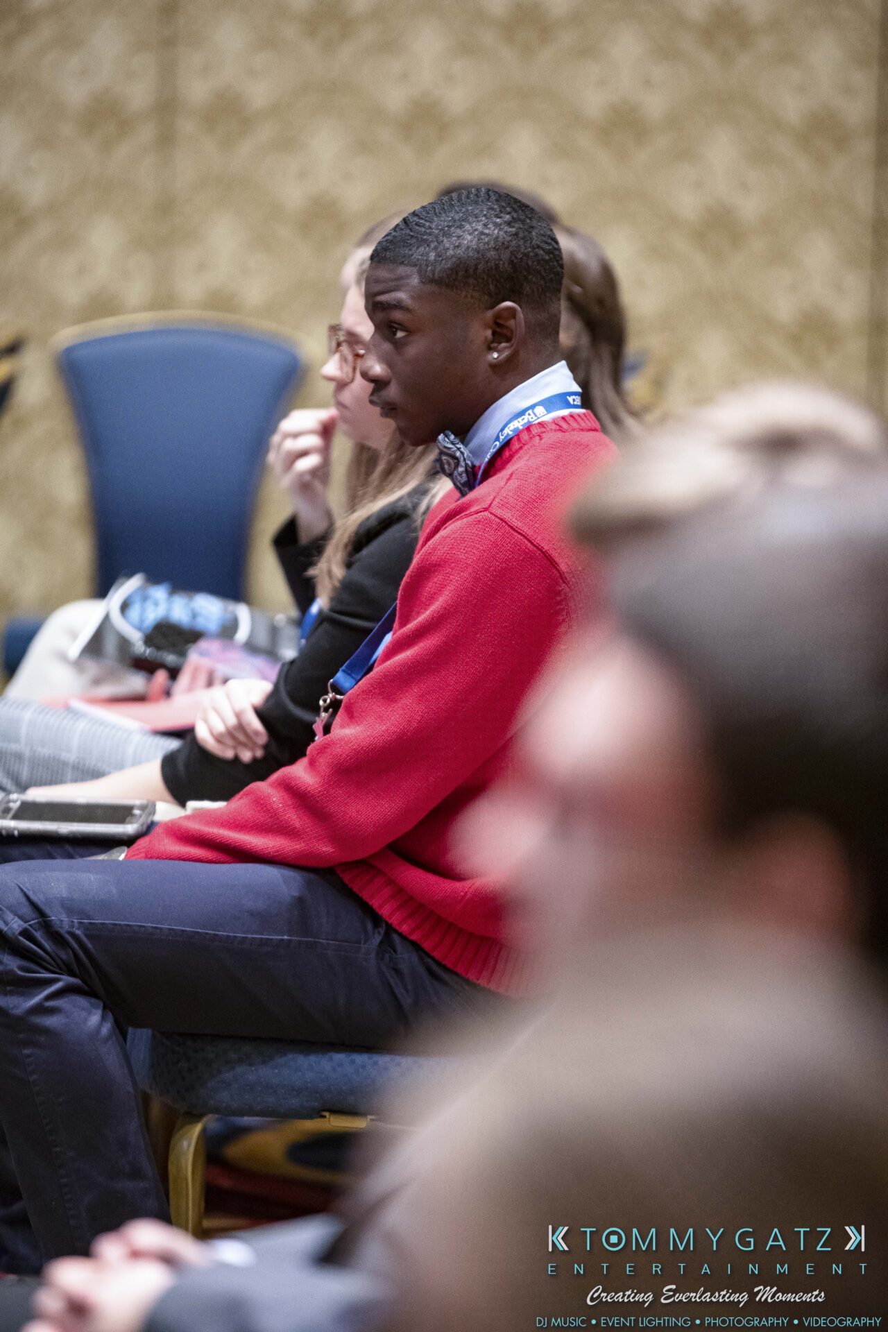 DECA-2018-Low-Res-WM-12 - TOMMY GATZ ENTERTAINMENT People seated in rows at an event, with a focus on a person in a red sweater attentively looking forward.