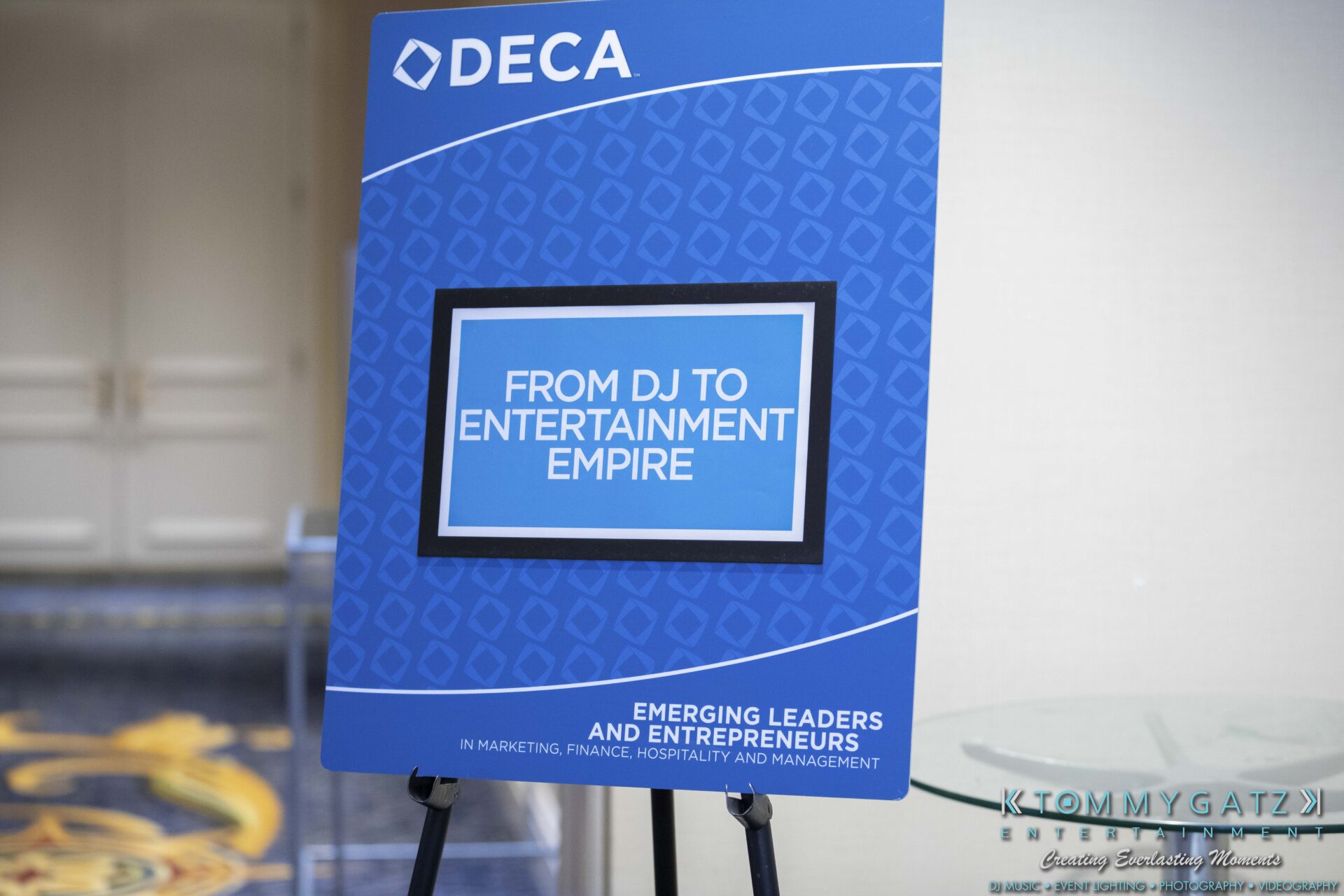 DECA-2018-Low-Res-WM-15 - TOMMY GATZ ENTERTAINMENT A blue sign with the DECA logo reads: "From DJ to Entertainment Empire. Emerging Leaders and Entrepreneurs in Marketing, Finance, Hospitality, and Management.