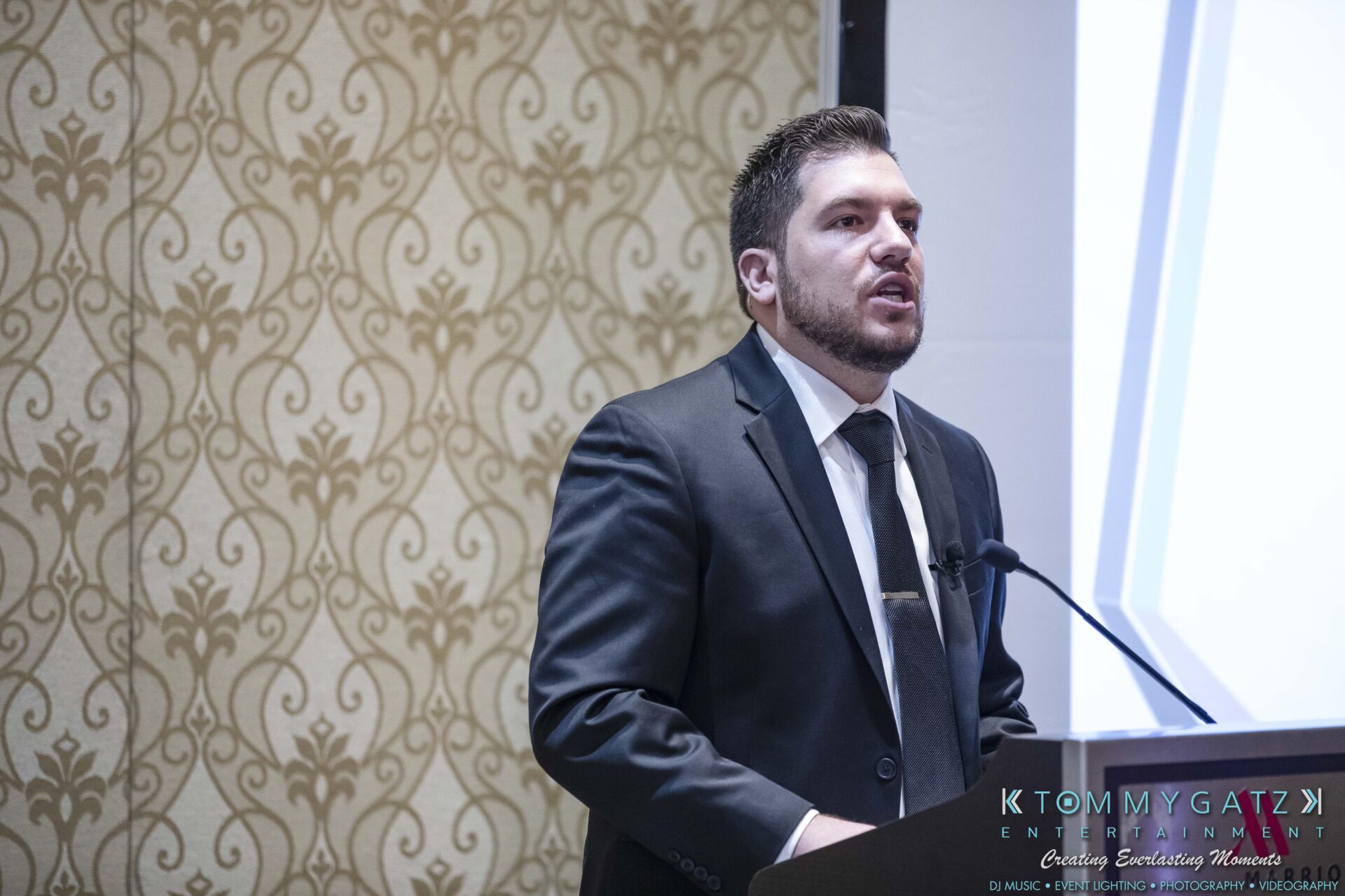 DECA-2018-Low-Res-WM-17 - TOMMY GATZ ENTERTAINMENT A man in a suit speaks at a podium in a formal setting with patterned wallpaper.