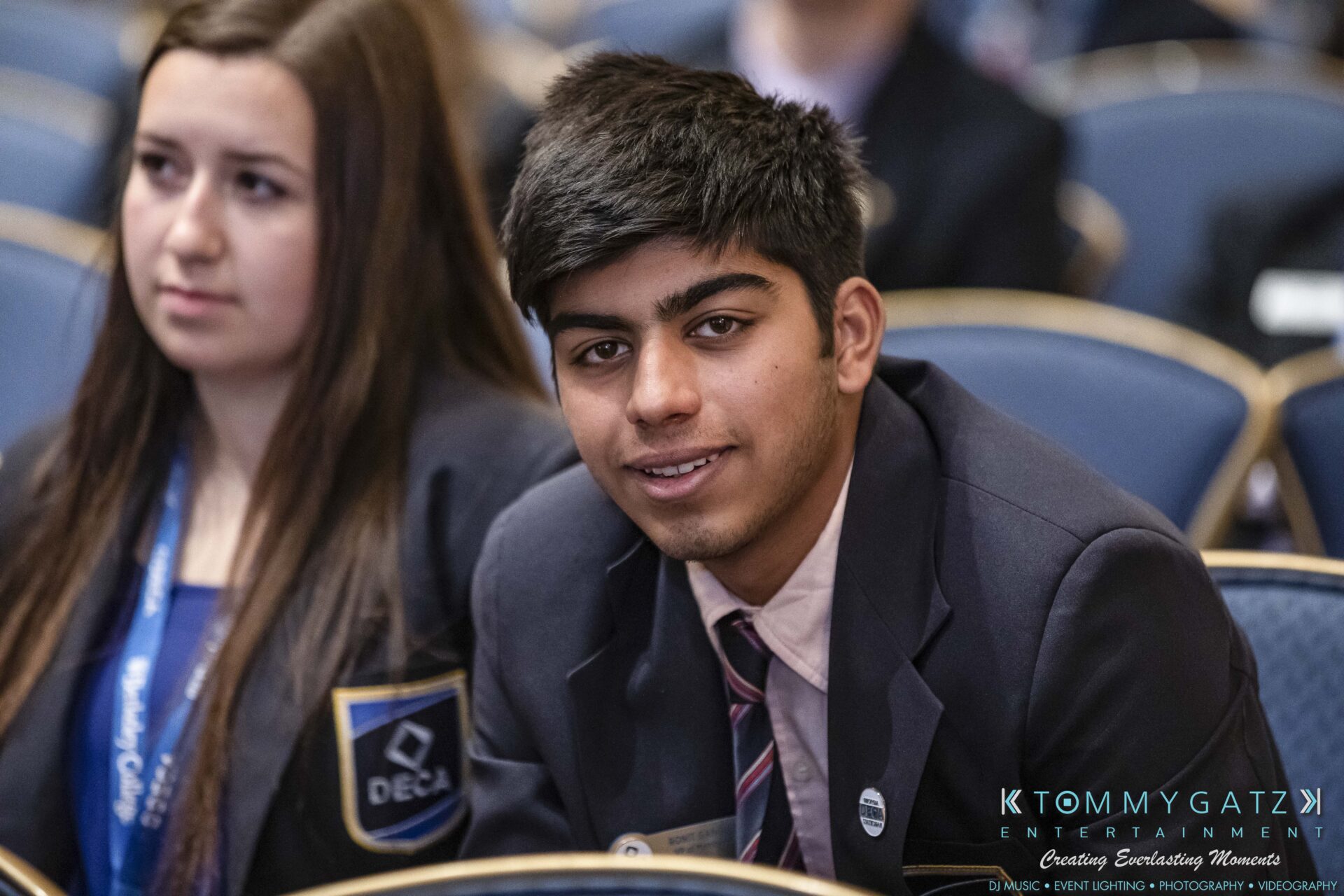 DECA-2018-Low-Res-WM-21 - TOMMY GATZ ENTERTAINMENT A young man and woman sit attentively in a row of blue chairs at an event, wearing formal attire.