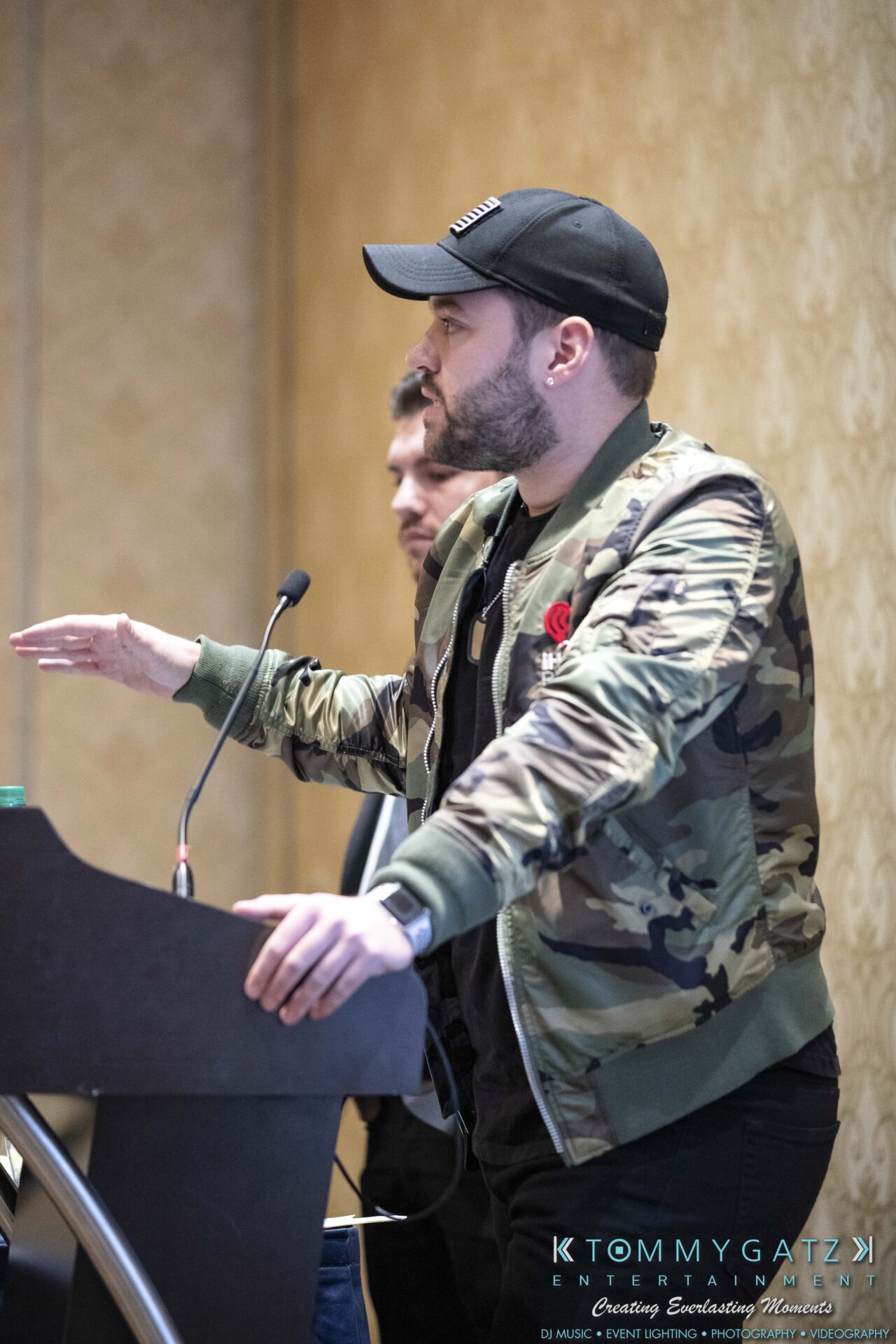 DECA-2018-Low-Res-WM-23 - TOMMY GATZ ENTERTAINMENT A man in a camo jacket and cap speaks at a podium with a microphone, gesturing with his hands.