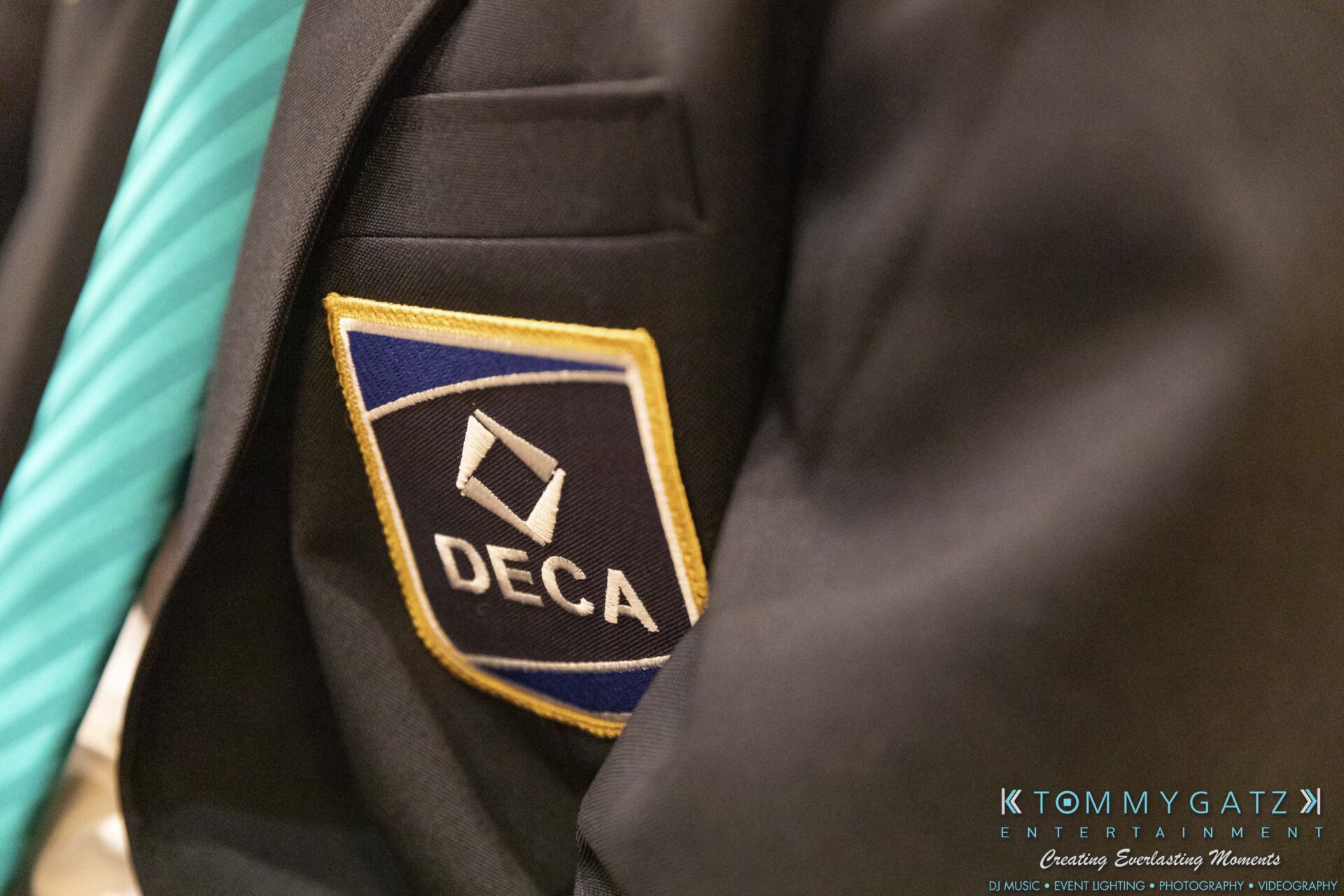 DECA-2018-Low-Res-WM-32 - TOMMY GATZ ENTERTAINMENT Close-up of a blazer with a DECA patch and a teal striped tie. The logo "KTOMMY GATZ ENTERTAINMENT" is visible in the corner.