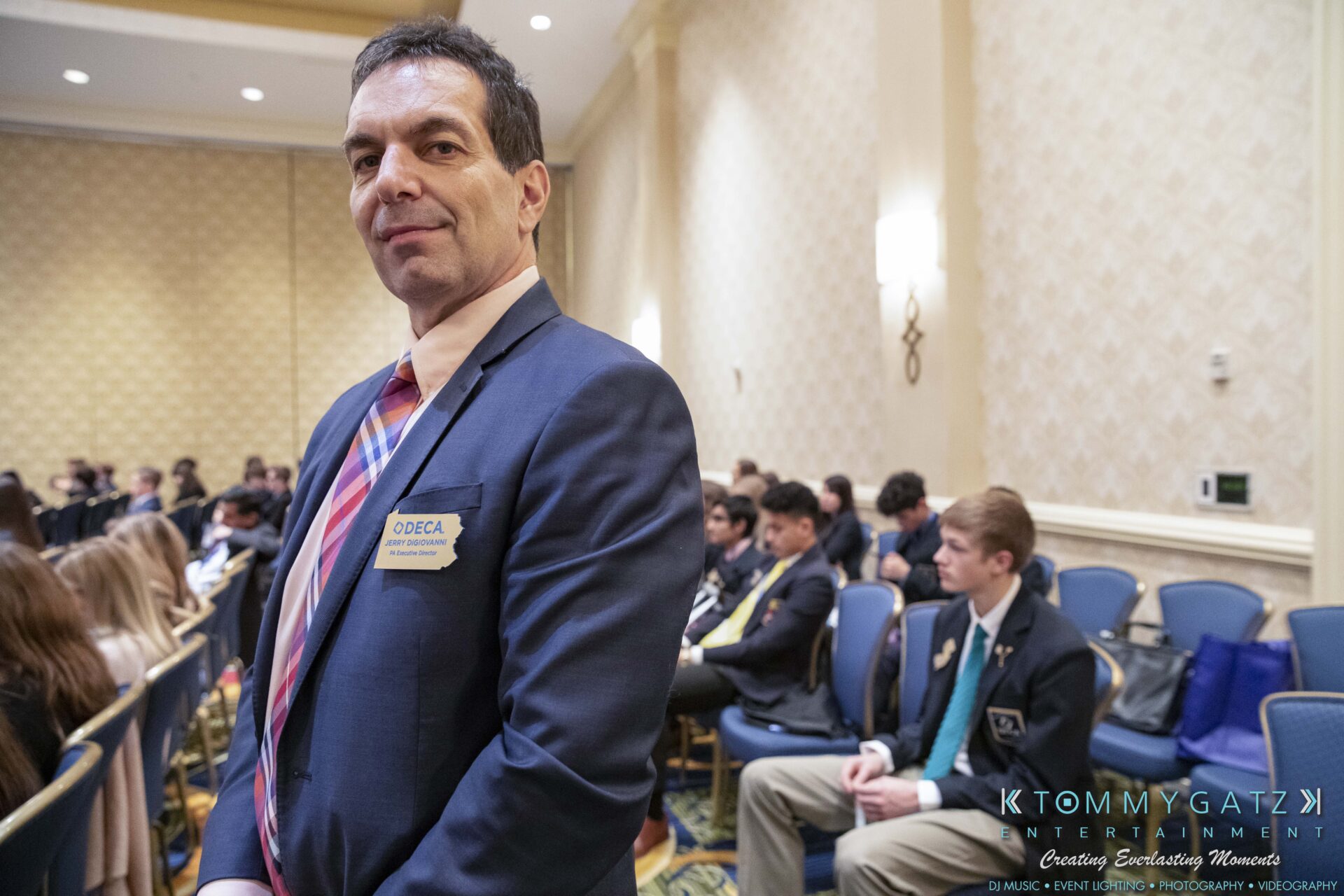 DECA-2018-Low-Res-WM-34 - TOMMY GATZ ENTERTAINMENT A man in a suit stands in a conference room with seated attendees in the background.