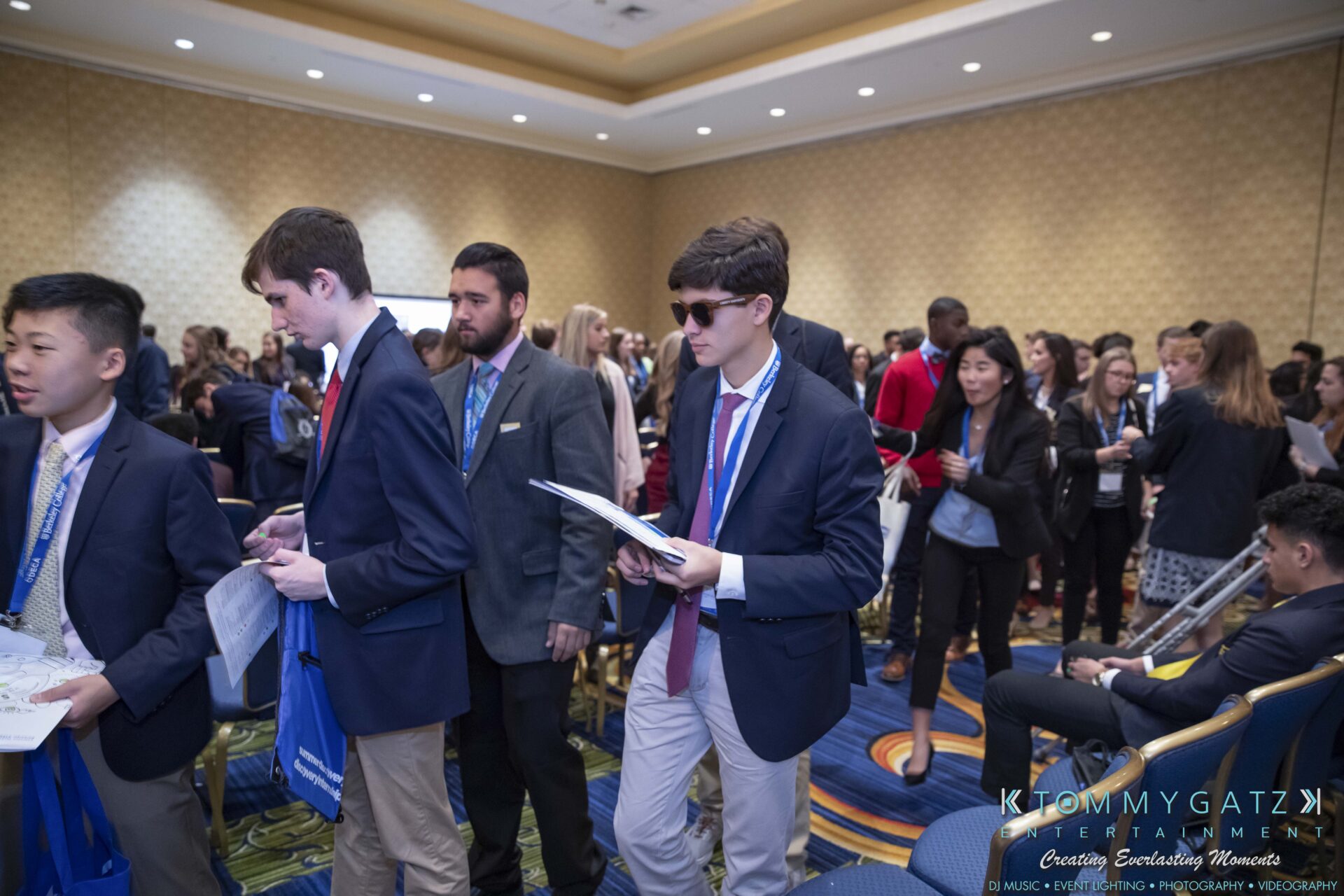 DECA-2018-Low-Res-WM-35 - TOMMY GATZ ENTERTAINMENT A crowd of young professionals in business attire walk through a conference room, holding papers and engaging with each other.