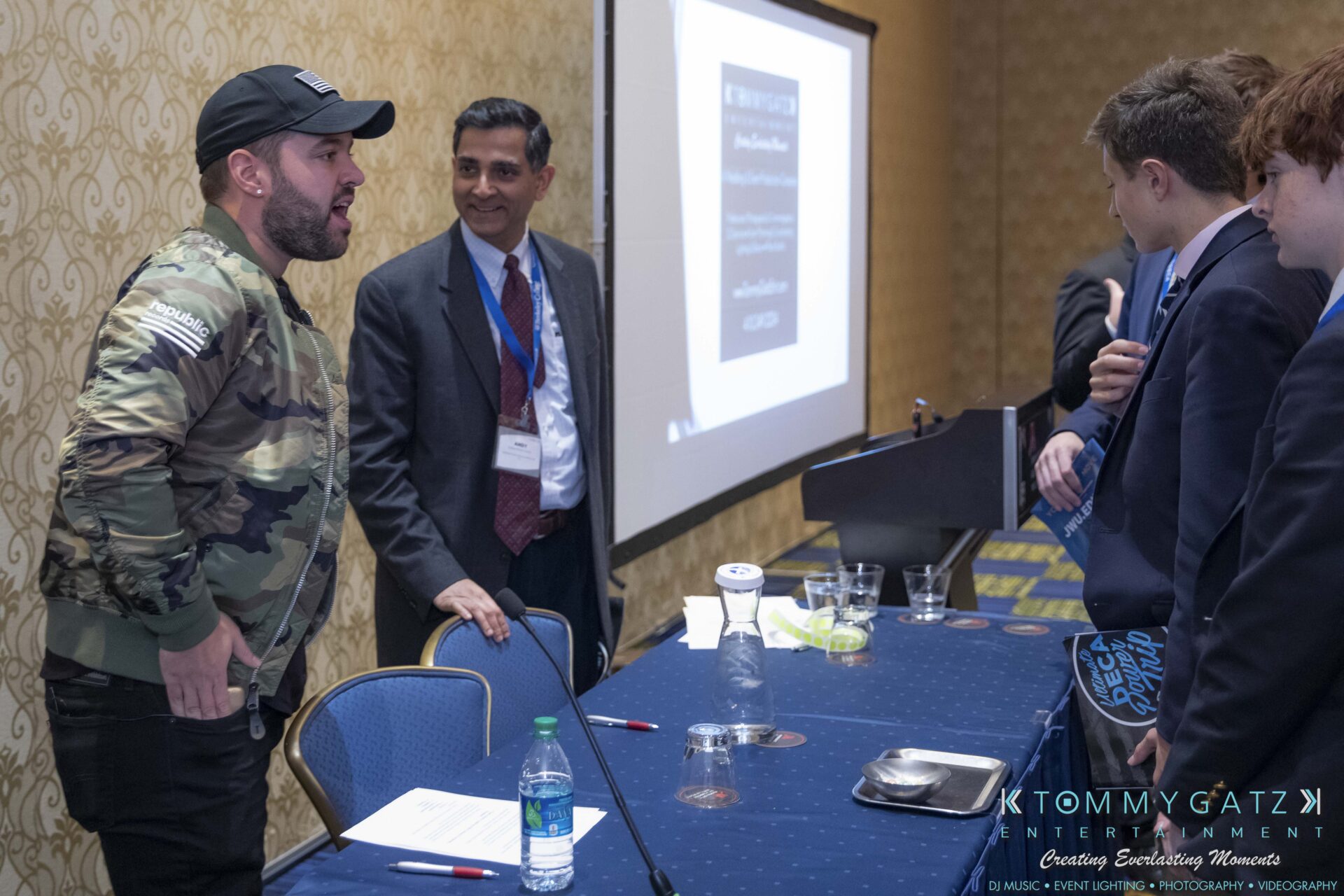 DECA-2018-Low-Res-WM-36 - TOMMY GATZ ENTERTAINMENT A man in a camo jacket speaks to a group of people around a table in a conference room. A screen displays indistinct text in the background.
