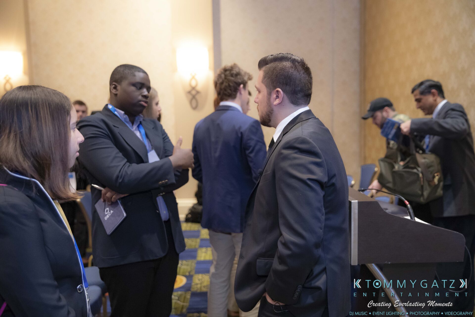 DECA-2018-Low-Res-WM-37 - TOMMY GATZ ENTERTAINMENT People in formal attire having a discussion in a conference room. Some are standing and others are sitting near a podium.