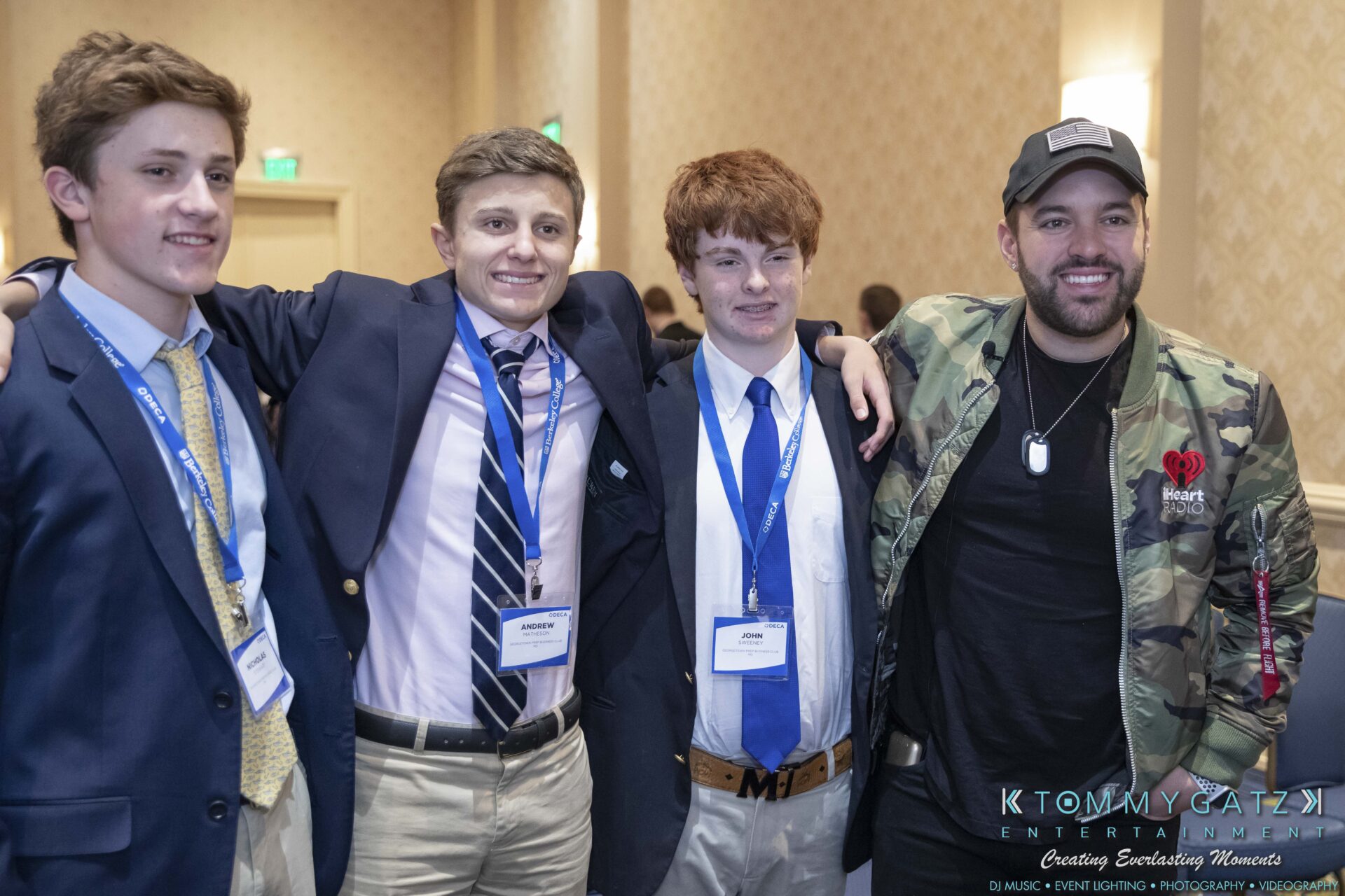 DECA-2018-Low-Res-WM-38 - TOMMY GATZ ENTERTAINMENT Four people pose together indoors. Three are wearing suits with name tags, and one is in a camouflage jacket and cap.