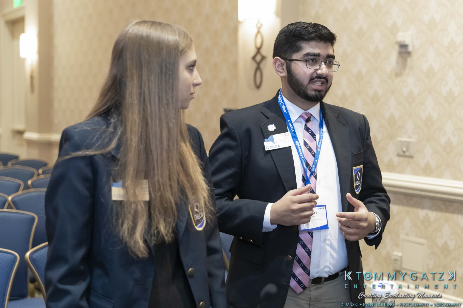 DECA-2018-Low-Res-WM-40 - TOMMY GATZ ENTERTAINMENT Two individuals in business attire are talking in a conference room. One person gestures with their hands, while the other listens. Both wear name tags and are engaged in conversation.