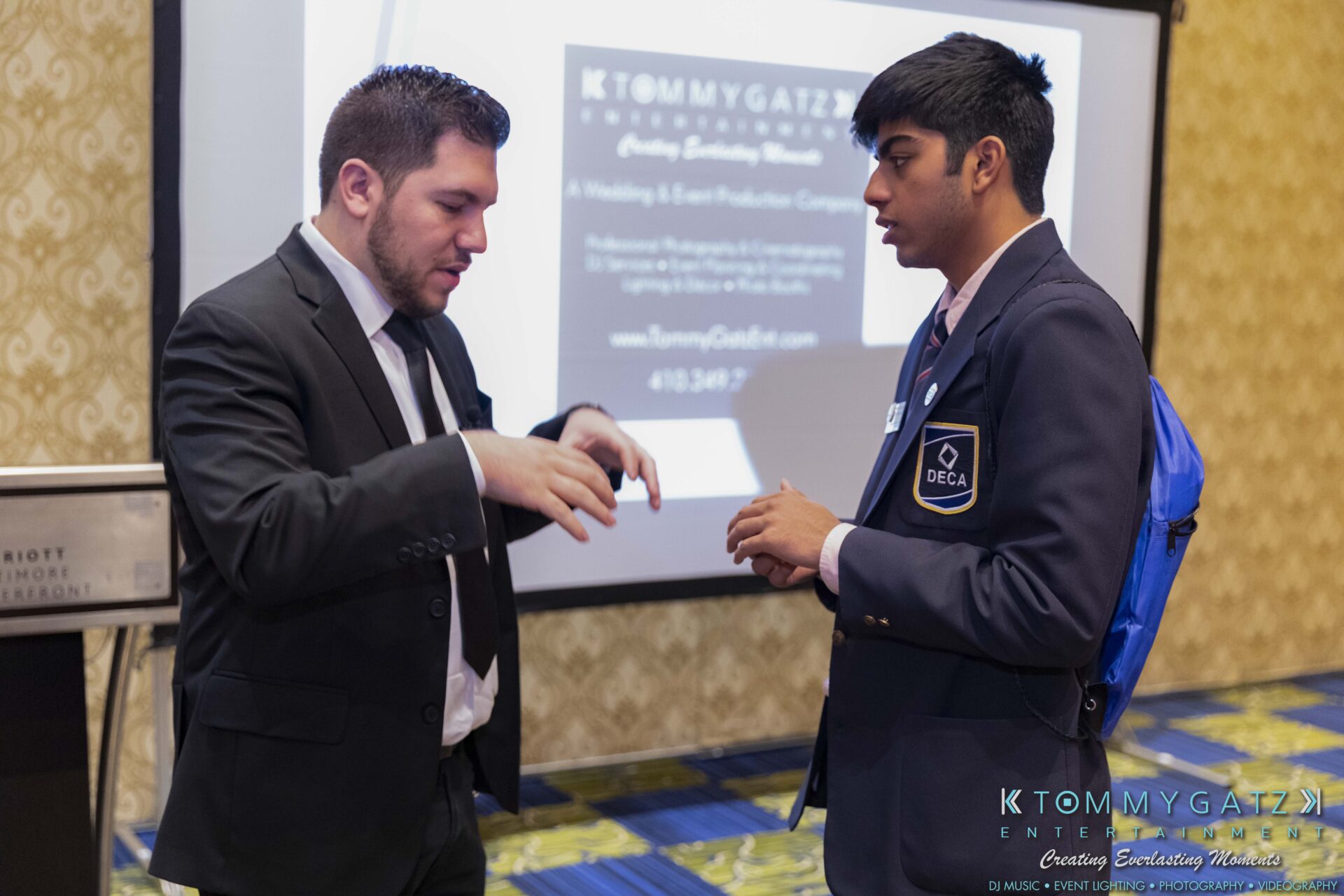 DECA-2018-Low-Res-WM-46 - TOMMY GATZ ENTERTAINMENT Two men in business attire have a discussion in front of a presentation screen at an event. One wears a name badge and a DECA blazer.