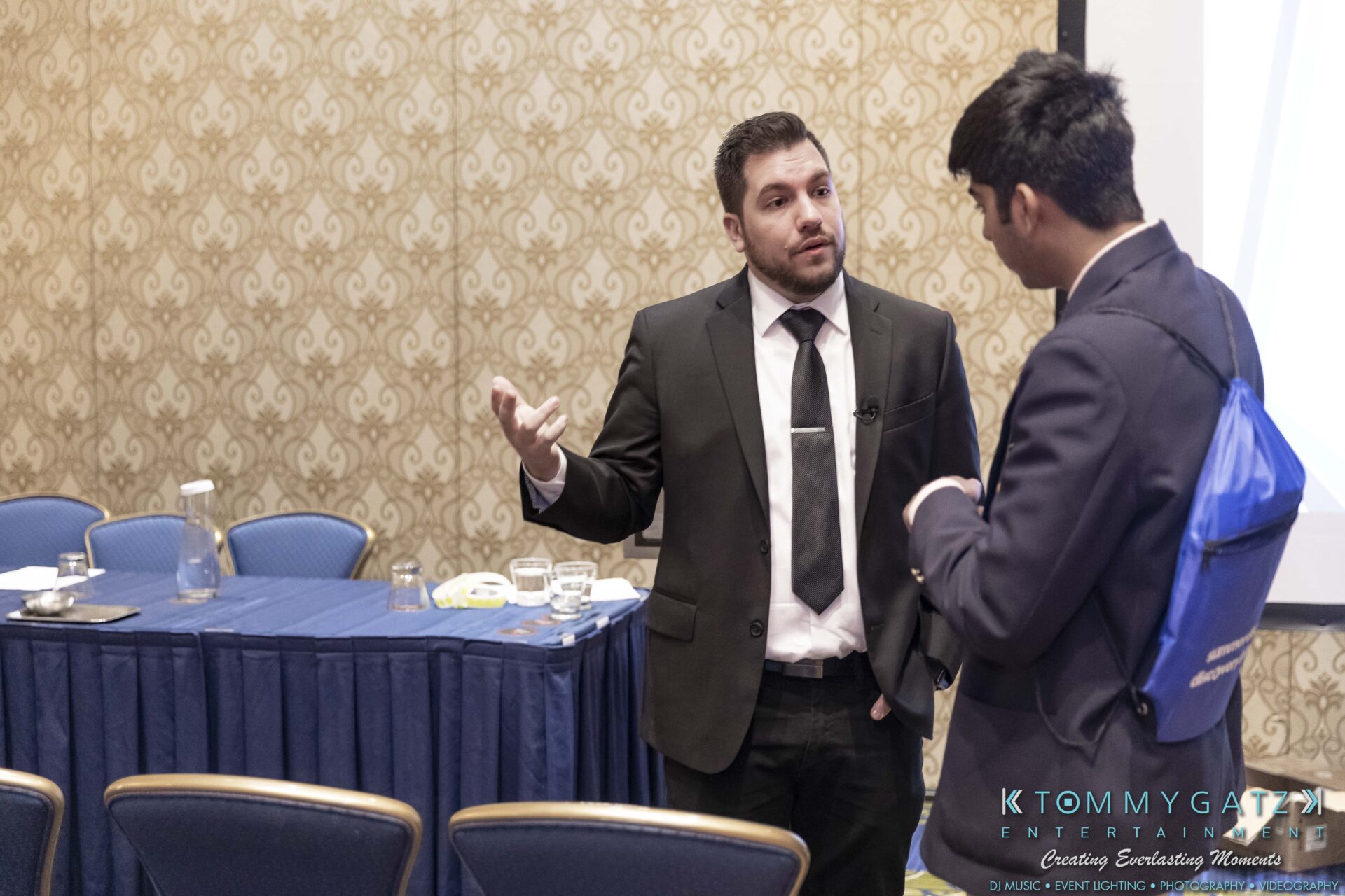 DECA-2018-Low-Res-WM-47 - TOMMY GATZ ENTERTAINMENT Two men in suits engaged in conversation beside a conference table with blue tablecloth and empty chairs.