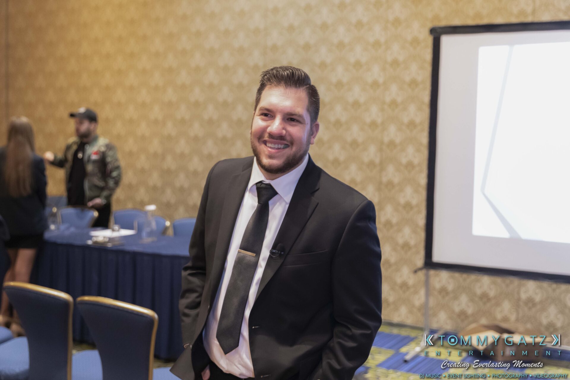 DECA-2018-Low-Res-WM-49 - TOMMY GATZ ENTERTAINMENT A man in a suit stands smiling in a conference room with a projection screen and several chairs.