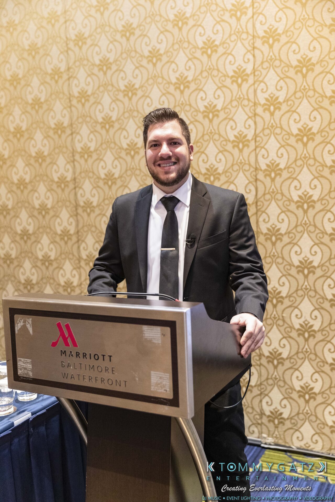 DECA-2018-Low-Res-WM-50 - TOMMY GATZ ENTERTAINMENT A man in a suit stands behind a podium in a conference room at the Marriott Baltimore Waterfront.