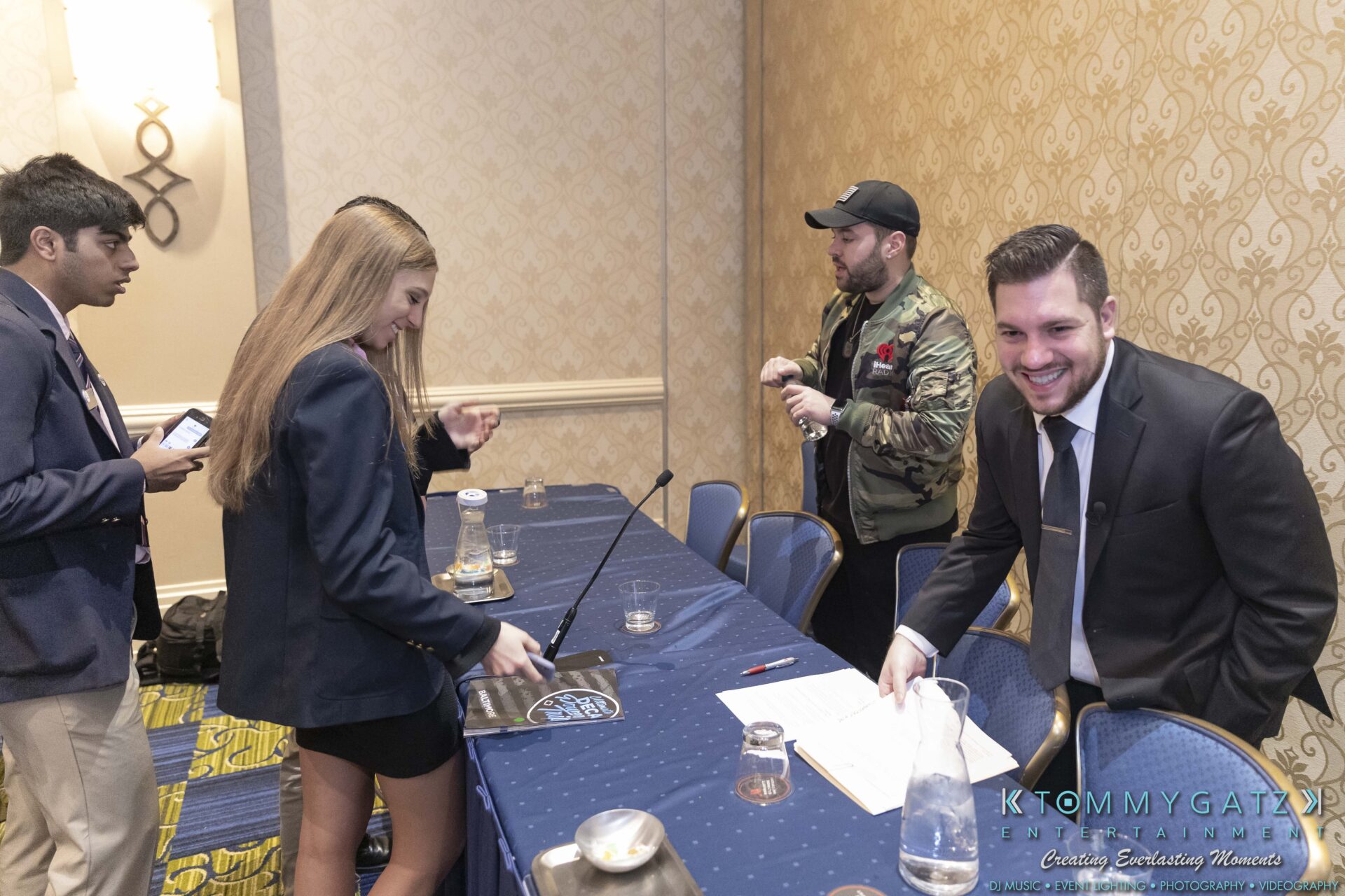 DECA-2018-Low-Res-WM-51 - TOMMY GATZ ENTERTAINMENT People gathered around a conference table, some standing and preparing to sit. A smiling man in a suit is in the foreground.