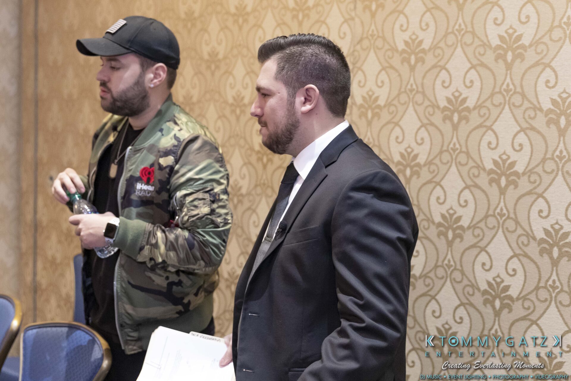 DECA-2018-Low-Res-WM-53 - TOMMY GATZ ENTERTAINMENT Two men stand side by side in a room. One wears a black baseball cap and camouflage jacket, holding a water bottle. The other is in a suit, holding papers.
