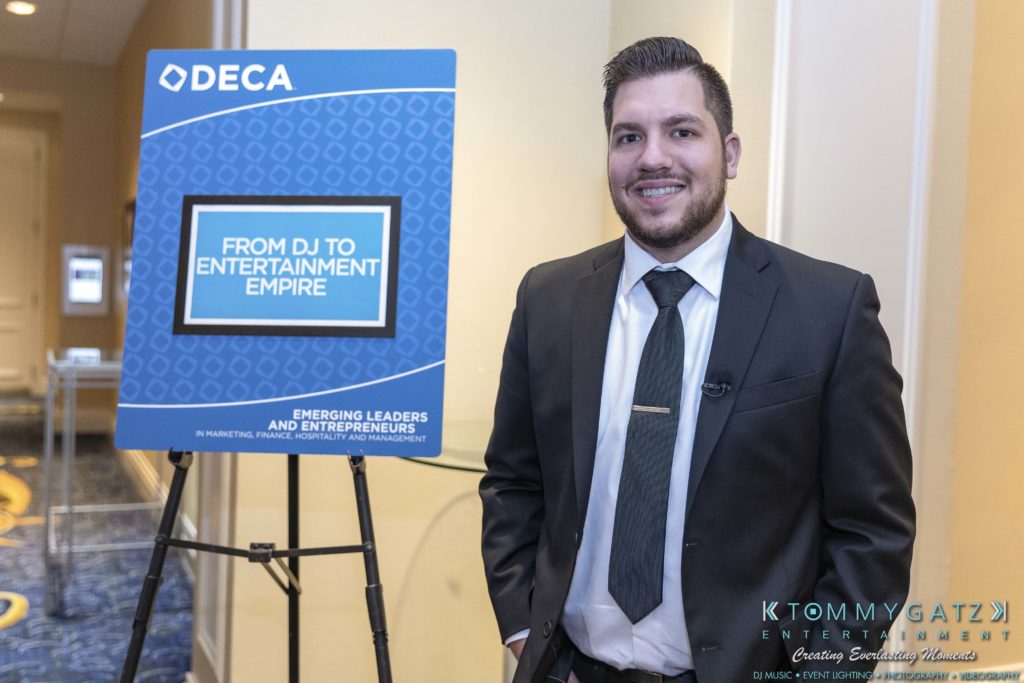 DECA-2018-Low-Res-WM-57-1024x683 - TOMMY GATZ ENTERTAINMENT A man in a suit stands by a sign that reads "From DJ to Entertainment Empire" at a DECA event.