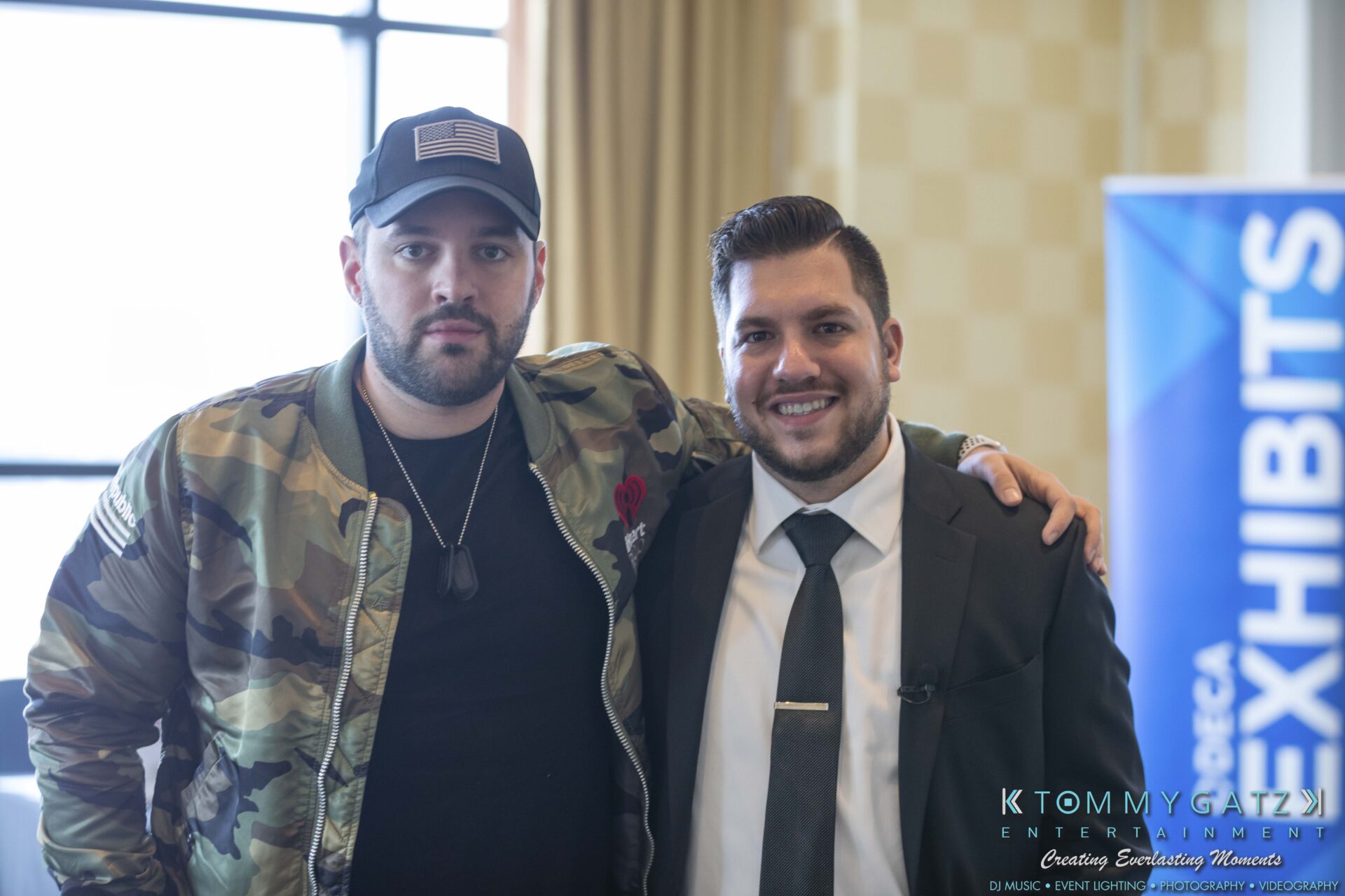 DECA-2018-Low-Res-WM-61 - TOMMY GATZ ENTERTAINMENT Two men are standing together, smiling. One is wearing a camo jacket and cap, and the other is in a suit and tie. A banner with the word "EXHIBITS" is partially visible in the background.