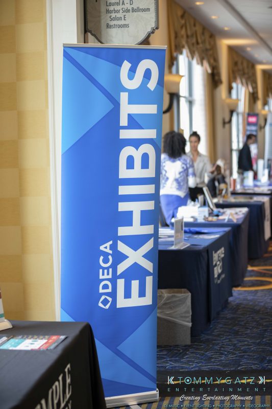 DECA-2018-Low-Res-WM-63-e1551129693180 - TOMMY GATZ ENTERTAINMENT A blue banner with the text "DECA Exhibits" stands in a hallway with people and tables exhibiting materials.