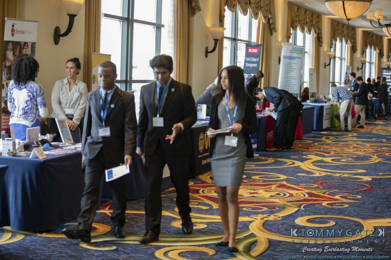 DECA-2018-Low-Res-WM-64-e1551129486651 - TOMMY GATZ ENTERTAINMENT People walking and talking at a business expo, with tables and banners in the background.